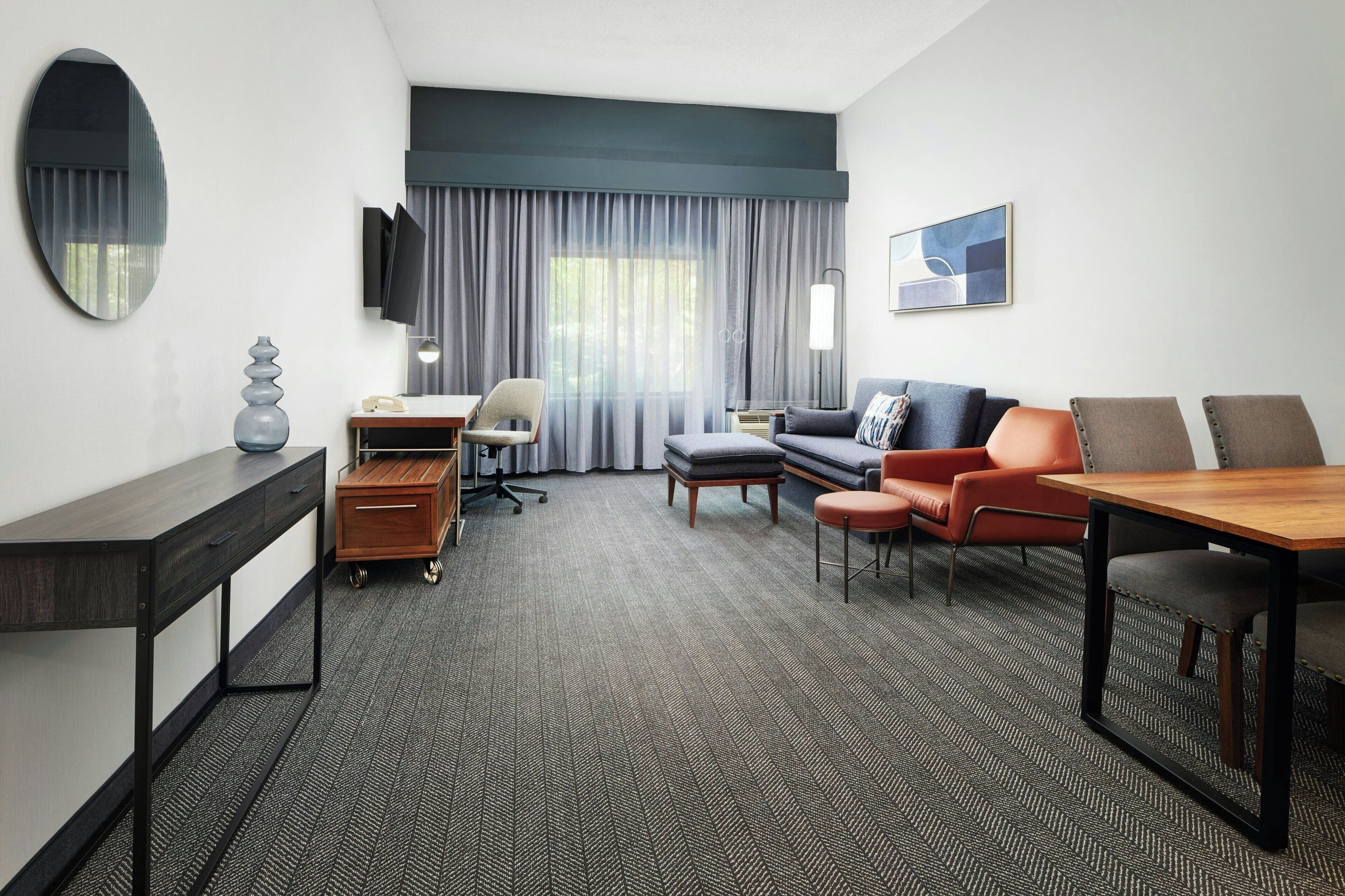 A hotel room with a sofa, armchairs, a dining table, a desk, a chair, a lamp, a TV, a window with curtains, and a painting on the wall.