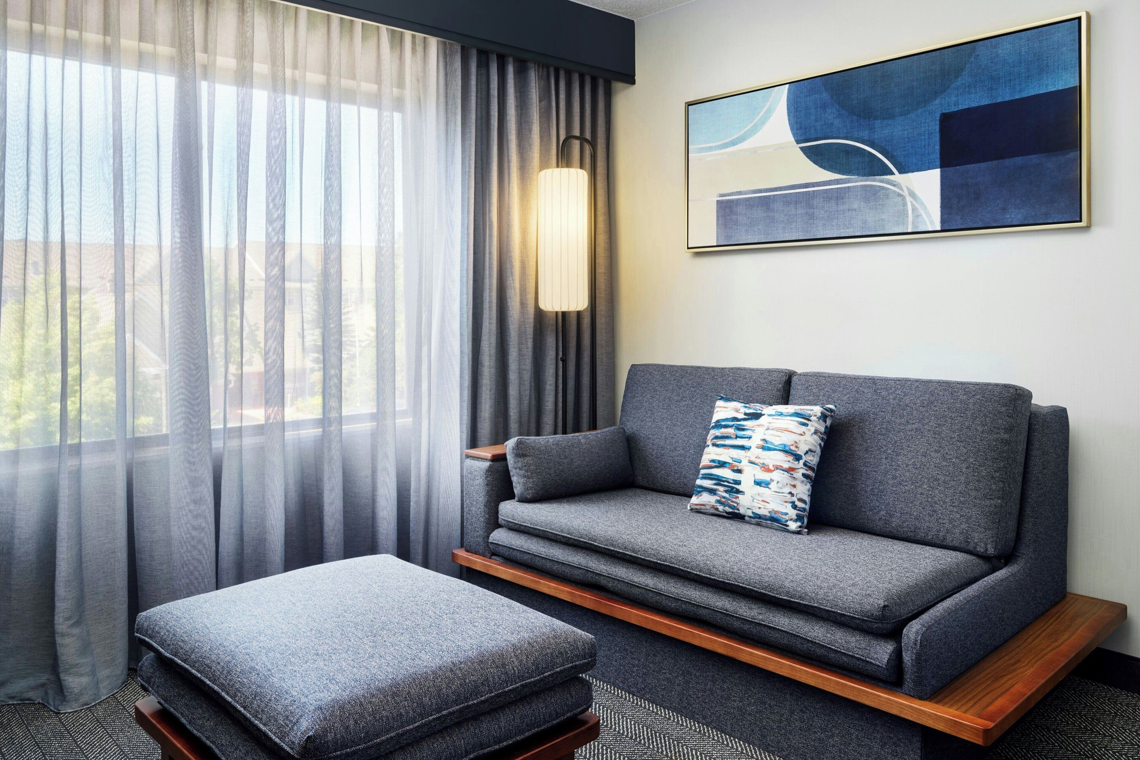 Un dormitorio de hotel moderno con un sofá gris, un reposapiés a juego, cortinas translúcidas, una lámpara de pie y una obra de arte abstracta en la pared.