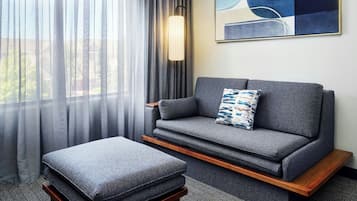 Un dormitorio de hotel moderno con un sofá gris, un reposapiés a juego, cortinas translúcidas, una lámpara de pie y una obra de arte abstracta en la pared.