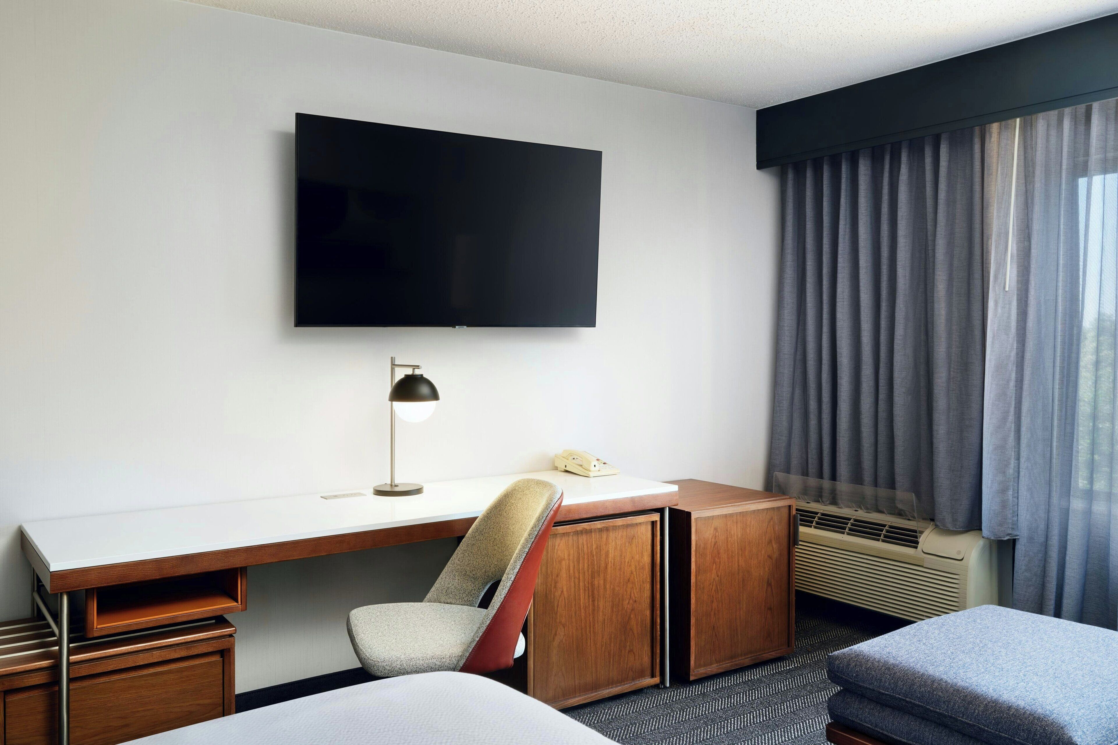 A hotel room with a flat-screen TV mounted on the wall, a desk with a chair, a telephone, and a window with curtains.