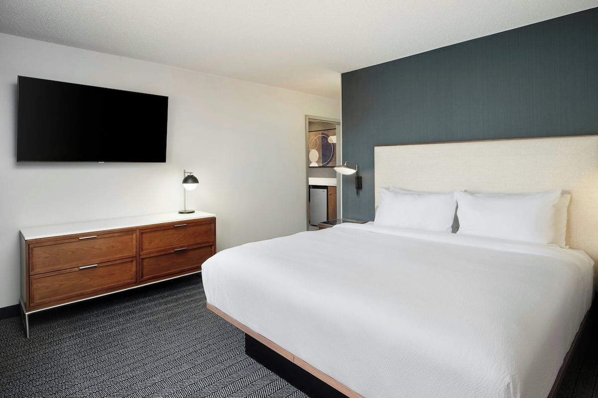 Quarto de hotel com uma cama grande, uma TV de tela plana na parede, uma cômoda de madeira e uma luminária de mesa.