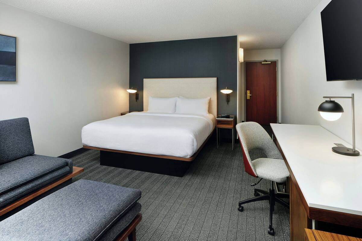 Quarto de hotel com cama, escrivaninha, cadeira e televisão fixada na parede.