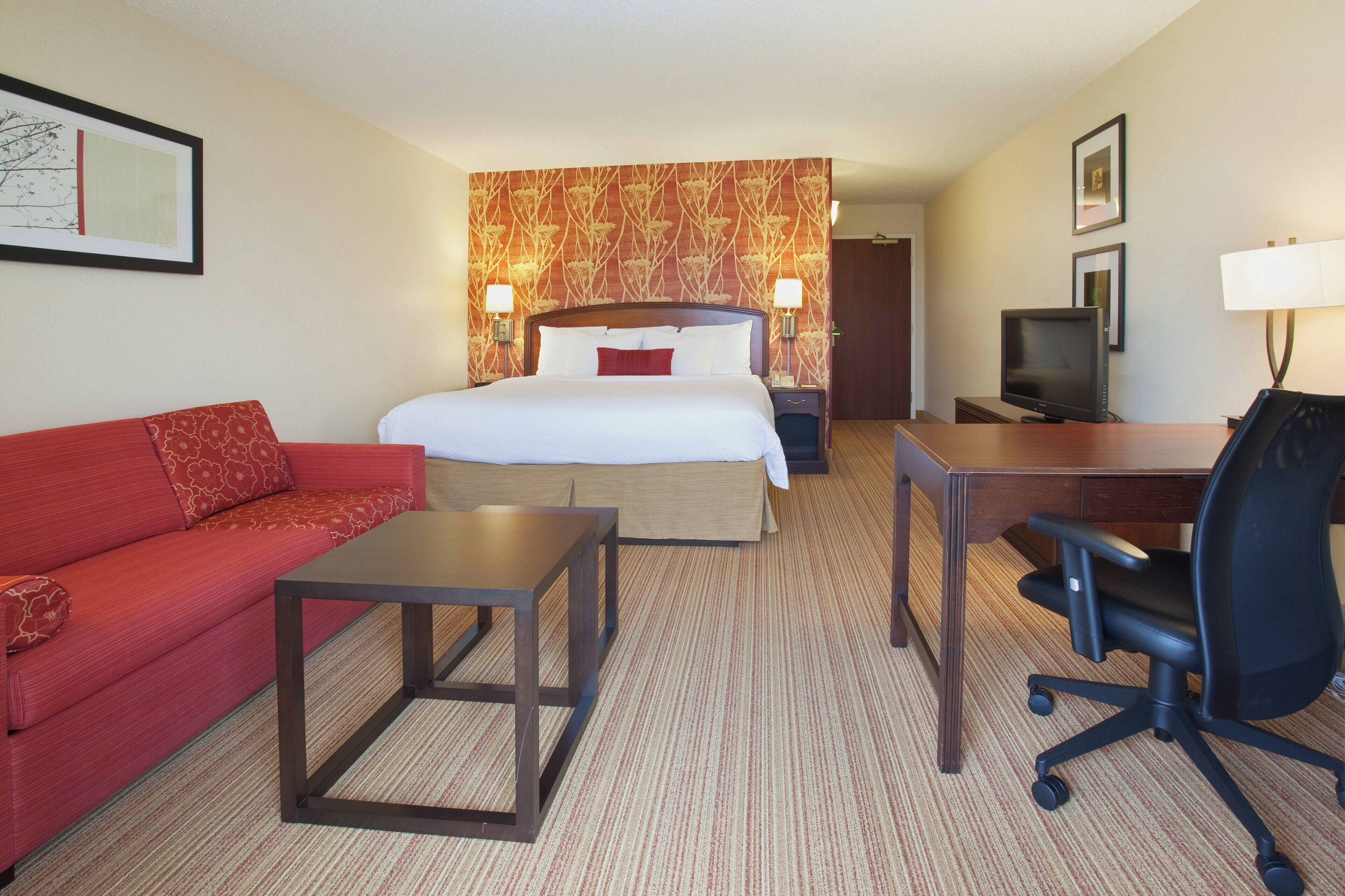 Habitación de hotel con cama, sofá rojo, escritorio con silla, televisor y lámpara.