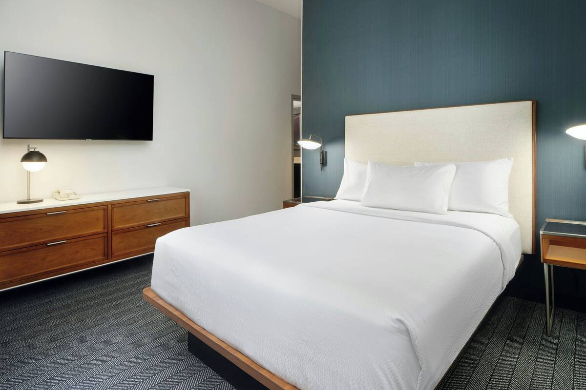 Quarto de hotel com uma cama grande, televisão de tela plana fixada na parede, cômoda de madeira e mesa de cabeceira com abajur.