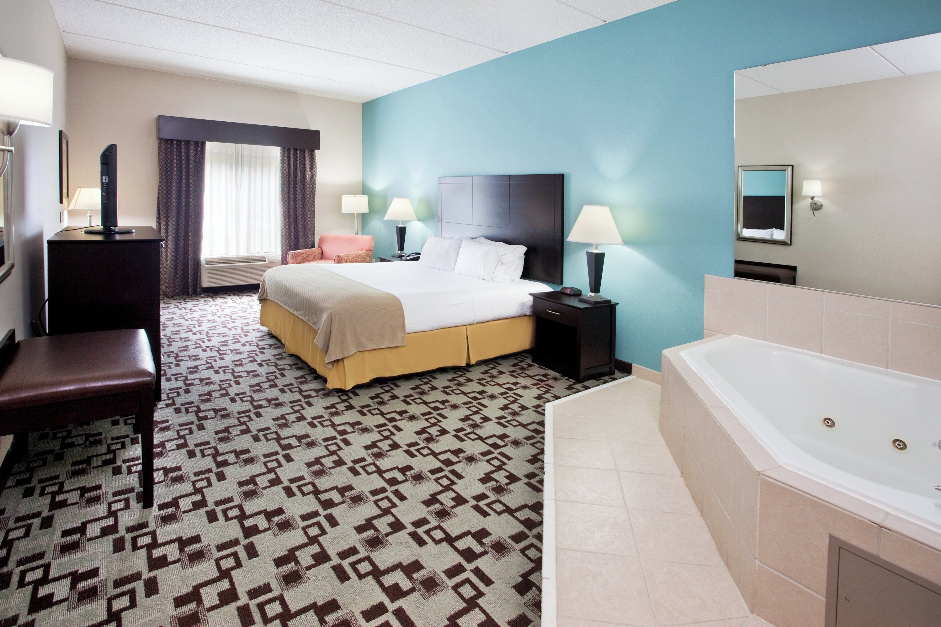 A hotel room with a bed, bedside tables, a desk, a chair, a bathroom with a bathtub, and a window with curtains.