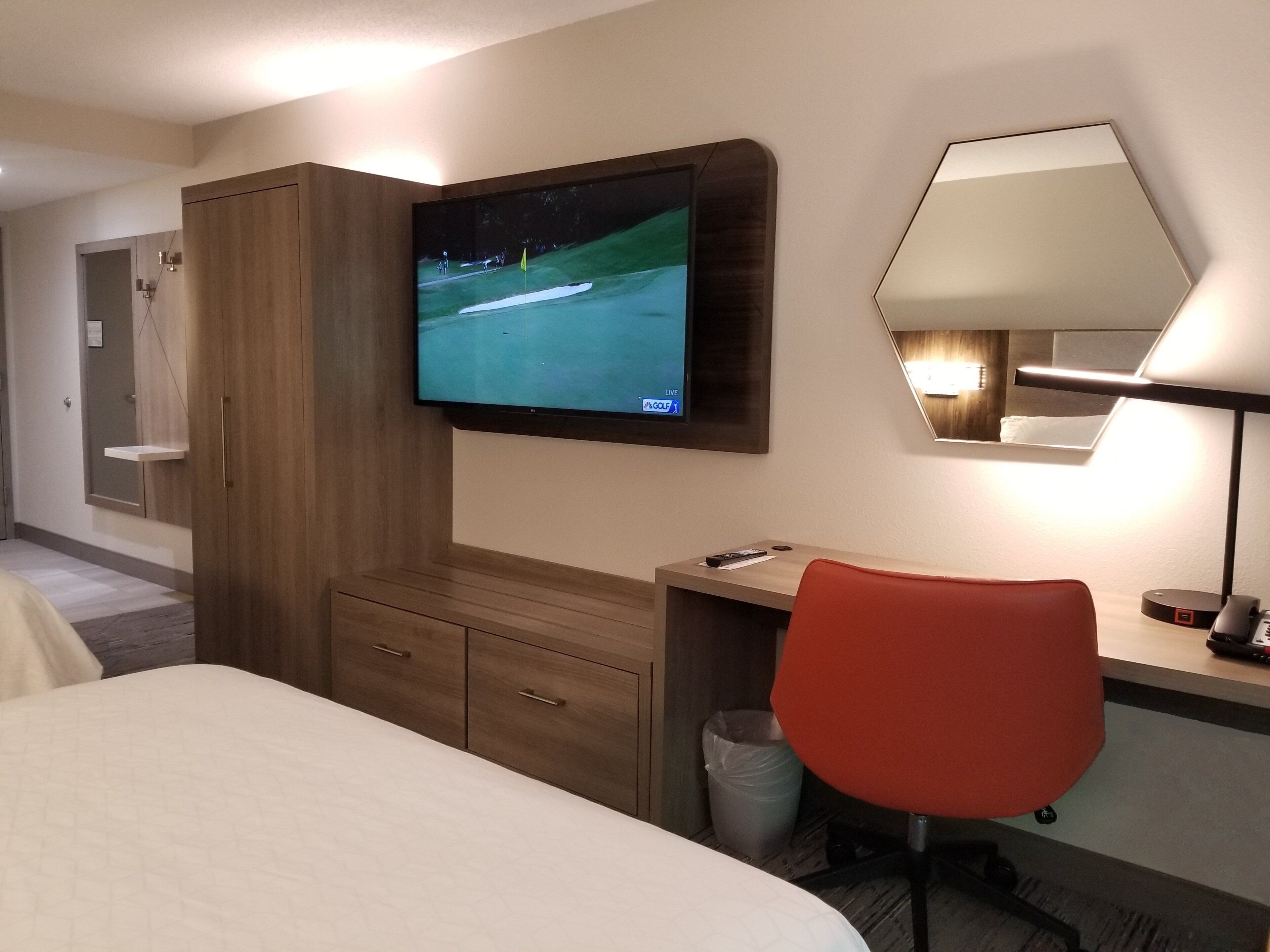 Une chambre d’hôtel comprenant un lit, un bureau avec une chaise, un téléviseur fixé au mur, un miroir et une lampe.