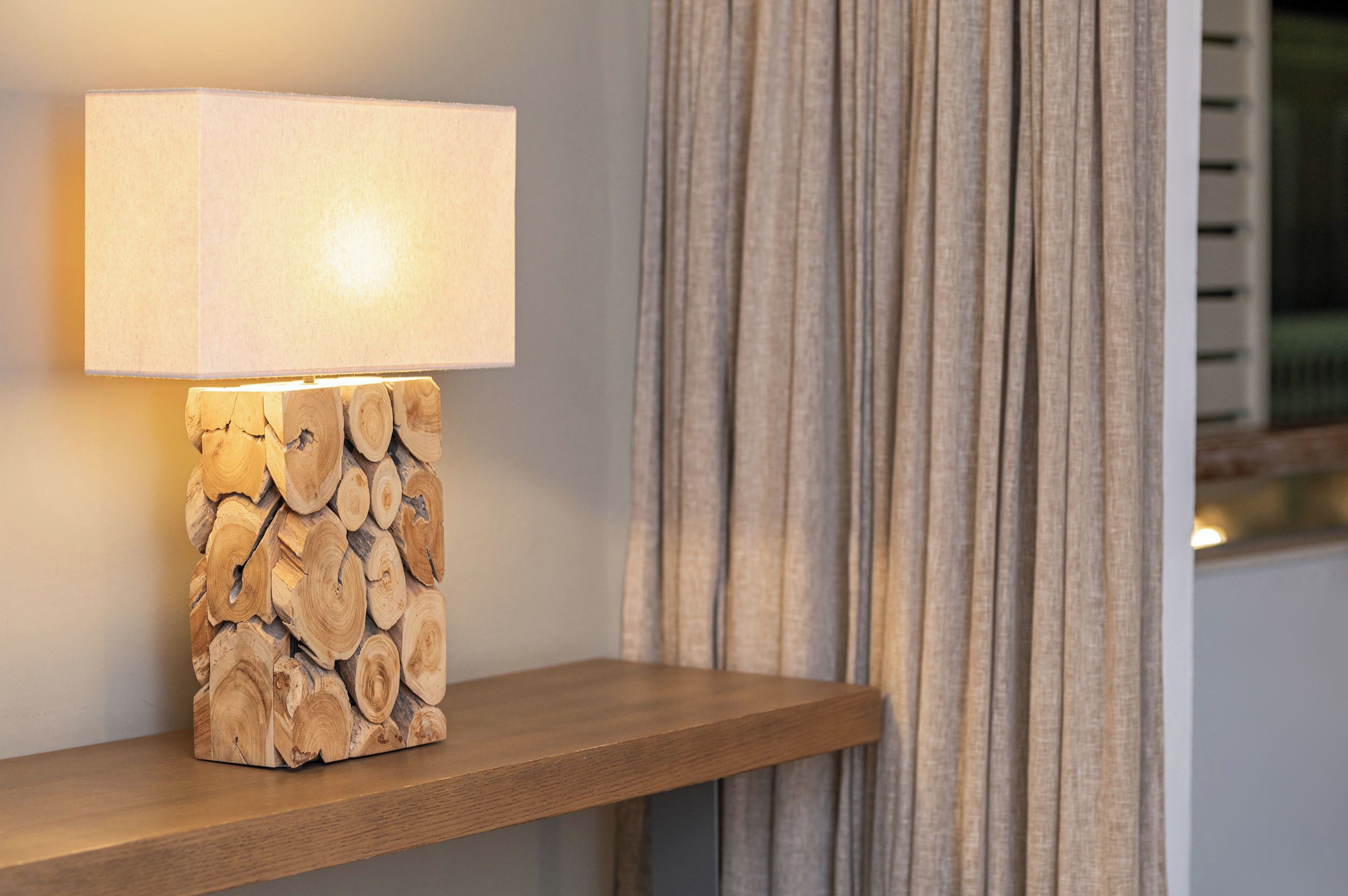 Eine Tischlampe mit hölzernem Fuß und weißem Lampenschirm, auf einem Holzbrett an einer Wand mit Vorhang.