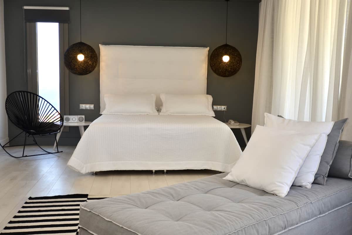 Un dormitorio moderno con una cama grande, un sofá gris, un sillón negro y dos lámparas colgantes.
