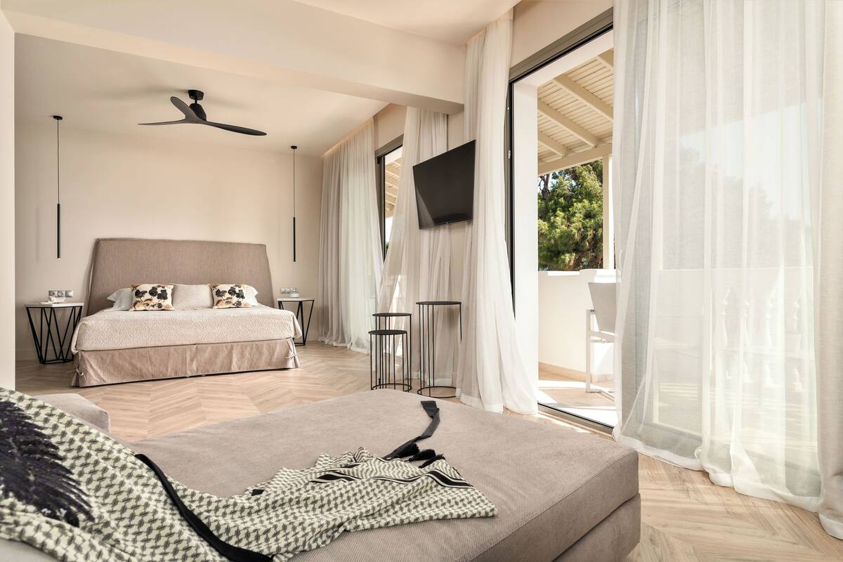 Un dormitorio moderno con una cama grande, ventilador de techo, televisor y balcón con cortinas traslúcidas.