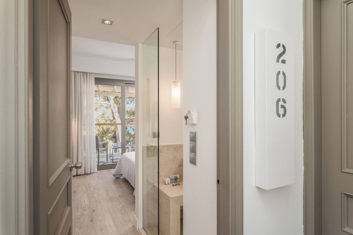 Un pasillo de hotel con una puerta de vidrio que da a un balcón con una cama. Se observa un baño con lavamanos y artículos de aseo. El número de habitación es 206.