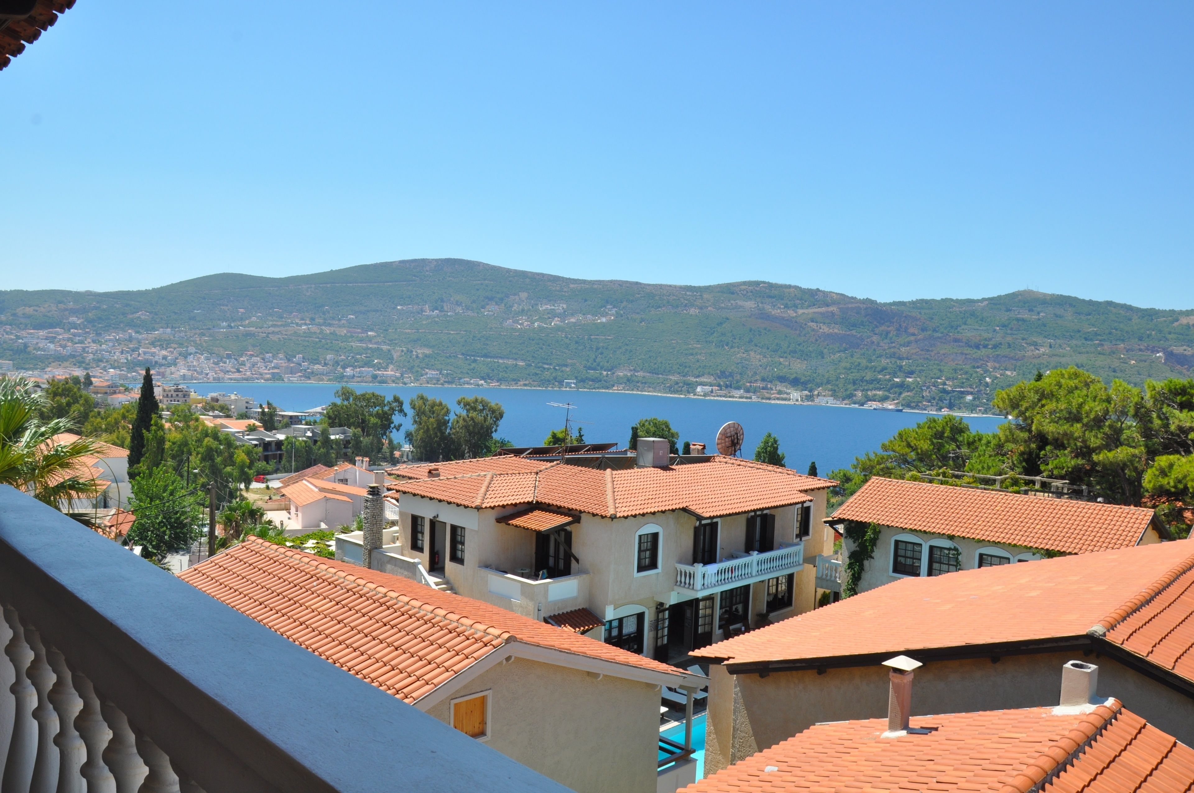 A coastal town with terracotta-roofed buildings, a clear blue sea, and green hills in the background.