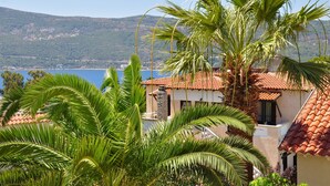 A coastal building with a red-tiled roof, surrounded by lush green palm trees and a view of the sea and hills in the background. - Ino Hotel & Suites (Samos)
