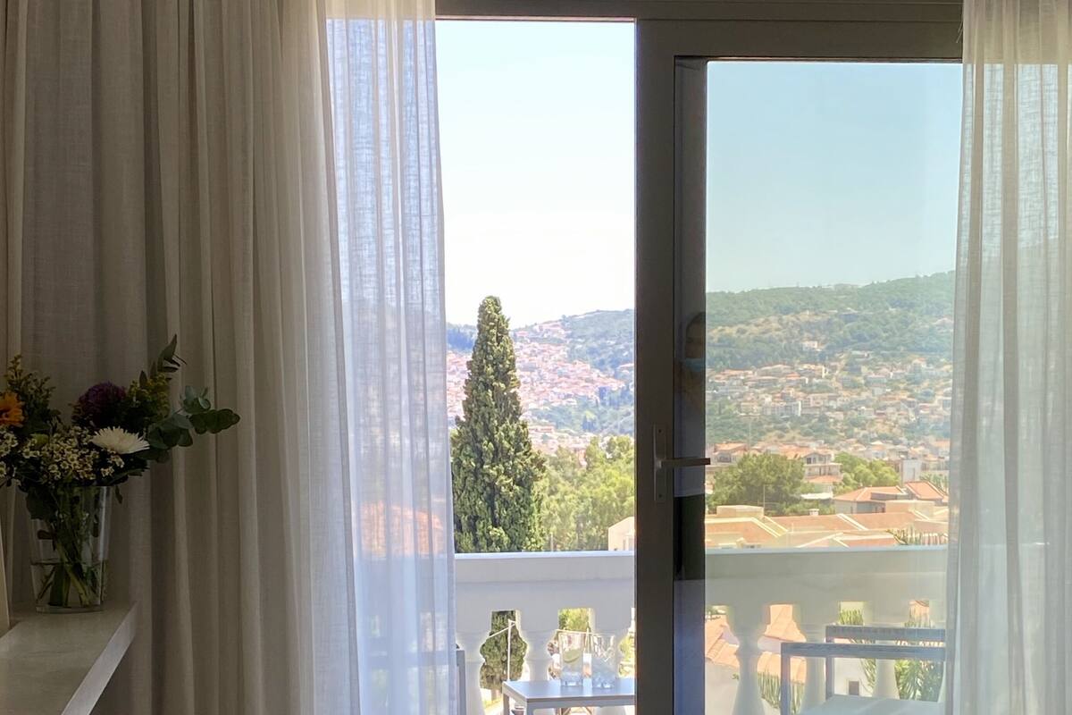 Un dormitorio con una cama grande, un balcón con mesa y sillas, y vistas a un pueblo en la ladera.