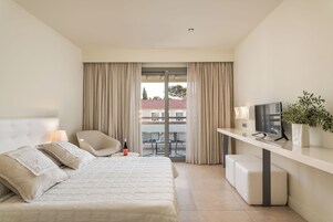 Habitación de hotel con cama, escritorio, silla, televisor y balcón con cortinas.