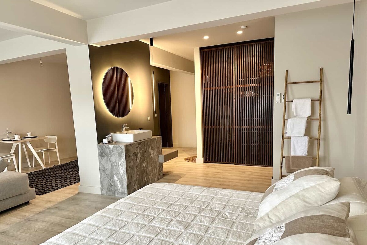 Een moderne slaapkamer met een groot bed, een badkamer met een wastafel en een zithoek met een tafel en stoelen.