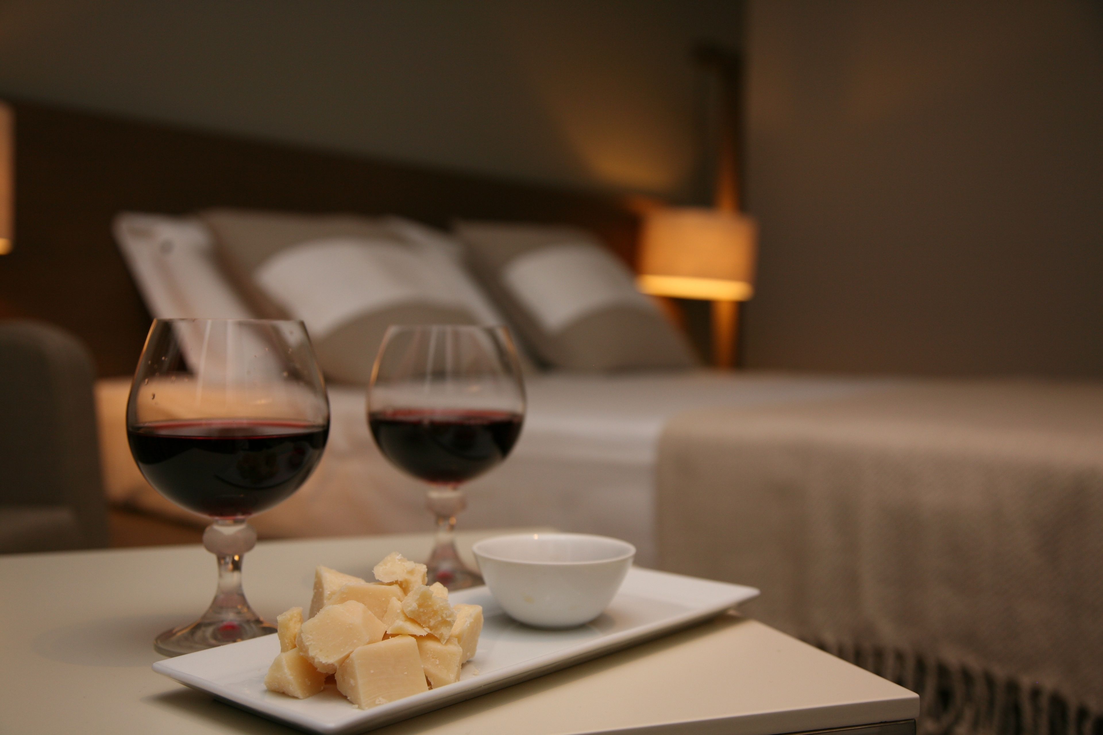 A neatly arranged table with two wine glasses, a bowl of cheese, and a bed in the background.