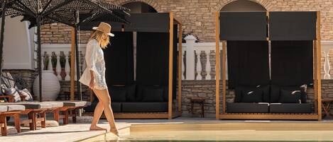 Zona de piscina con sillones de descanso, un espacio de estar cubierto y un muro de piedra de fondo.