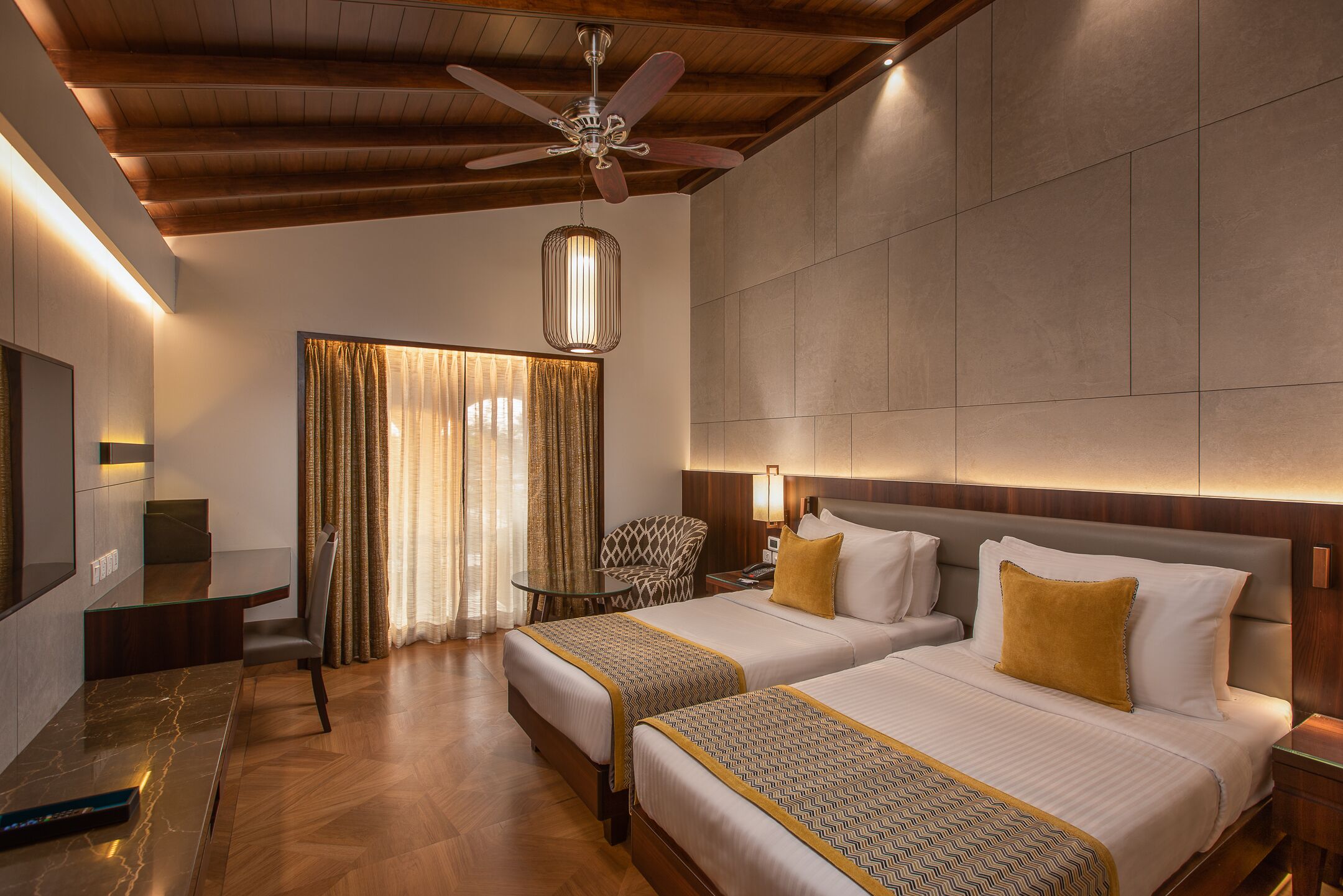 A hotel room with two beds, a desk, a chair, a ceiling fan, and a large window with curtains.