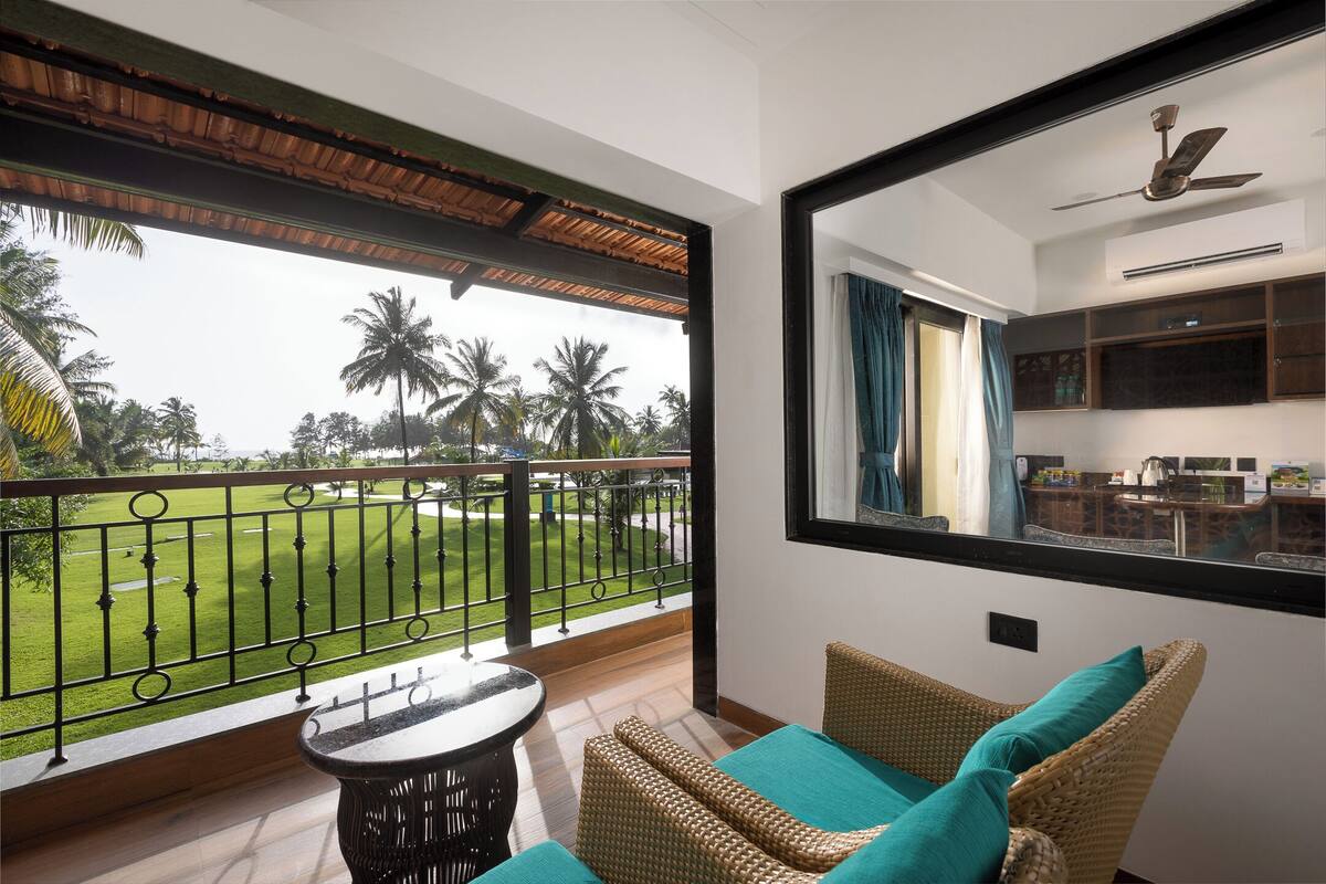 A balcony with a view of a well-maintained lawn and palm trees, a wicker chair with teal cushions, and a small round table.
