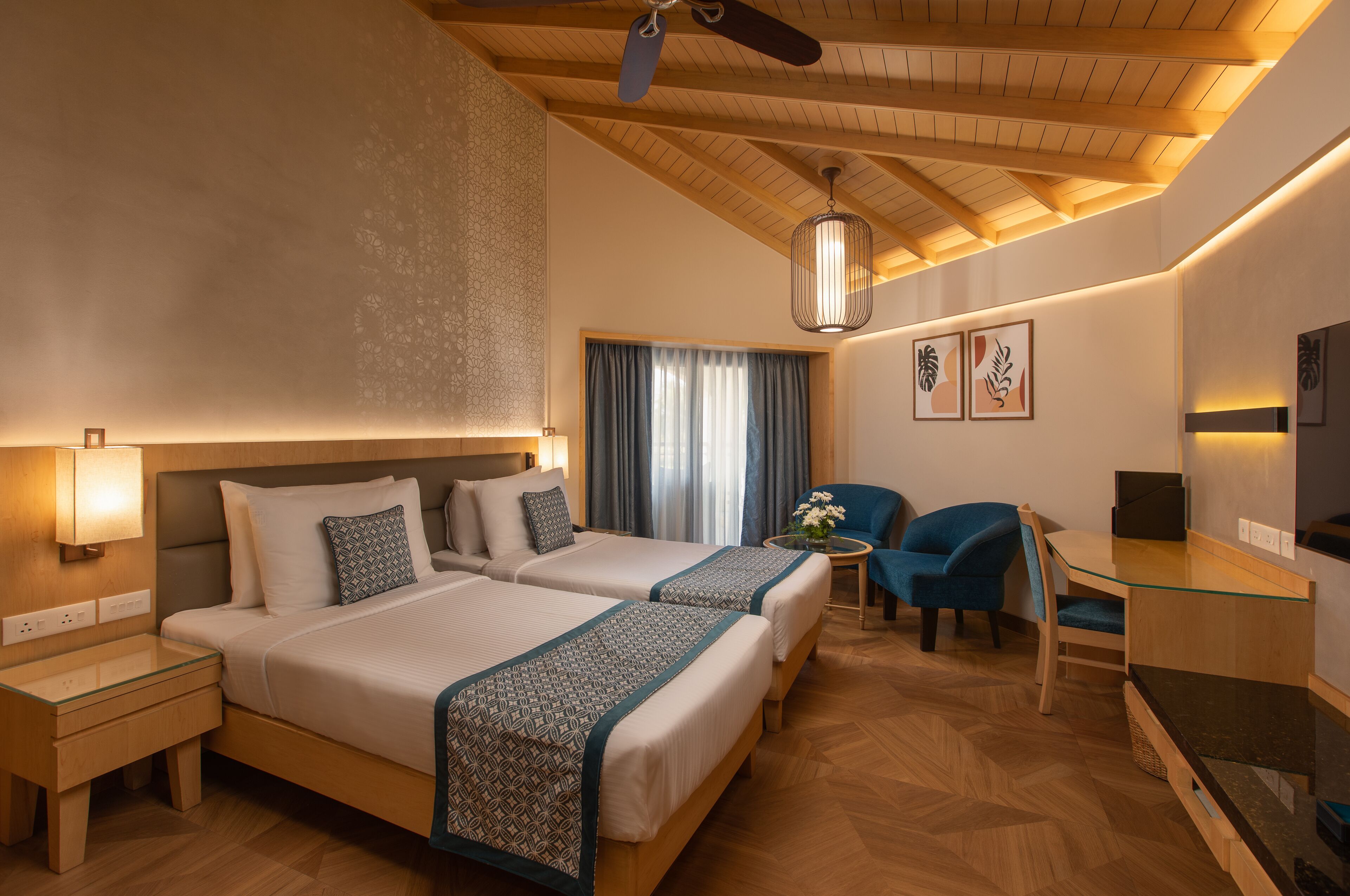 A hotel room with two beds, a desk, a chair, a ceiling fan, and a television.