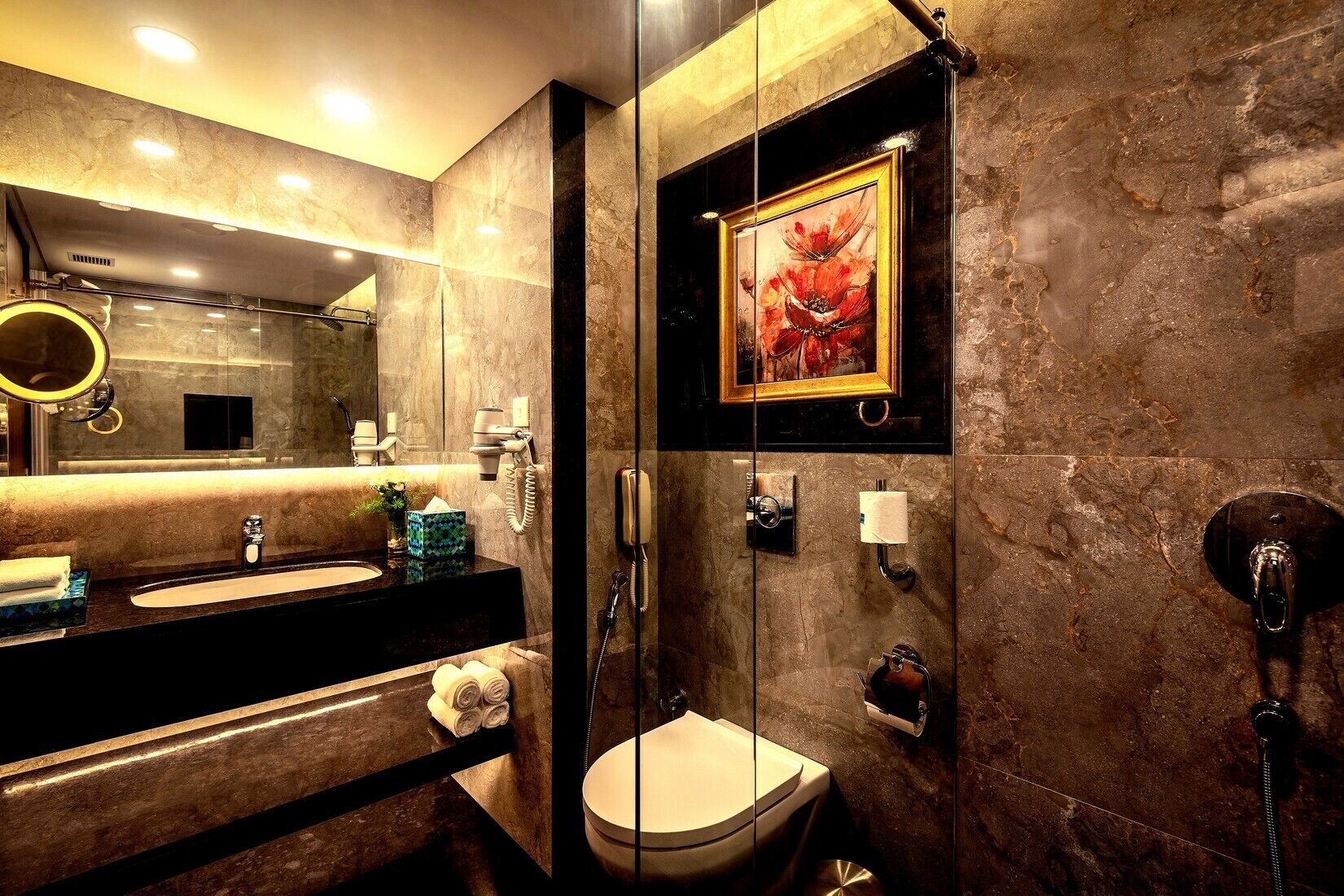 A modern bathroom with a marble wall, a large mirror, a sink with a black countertop, a toilet, and a shower.