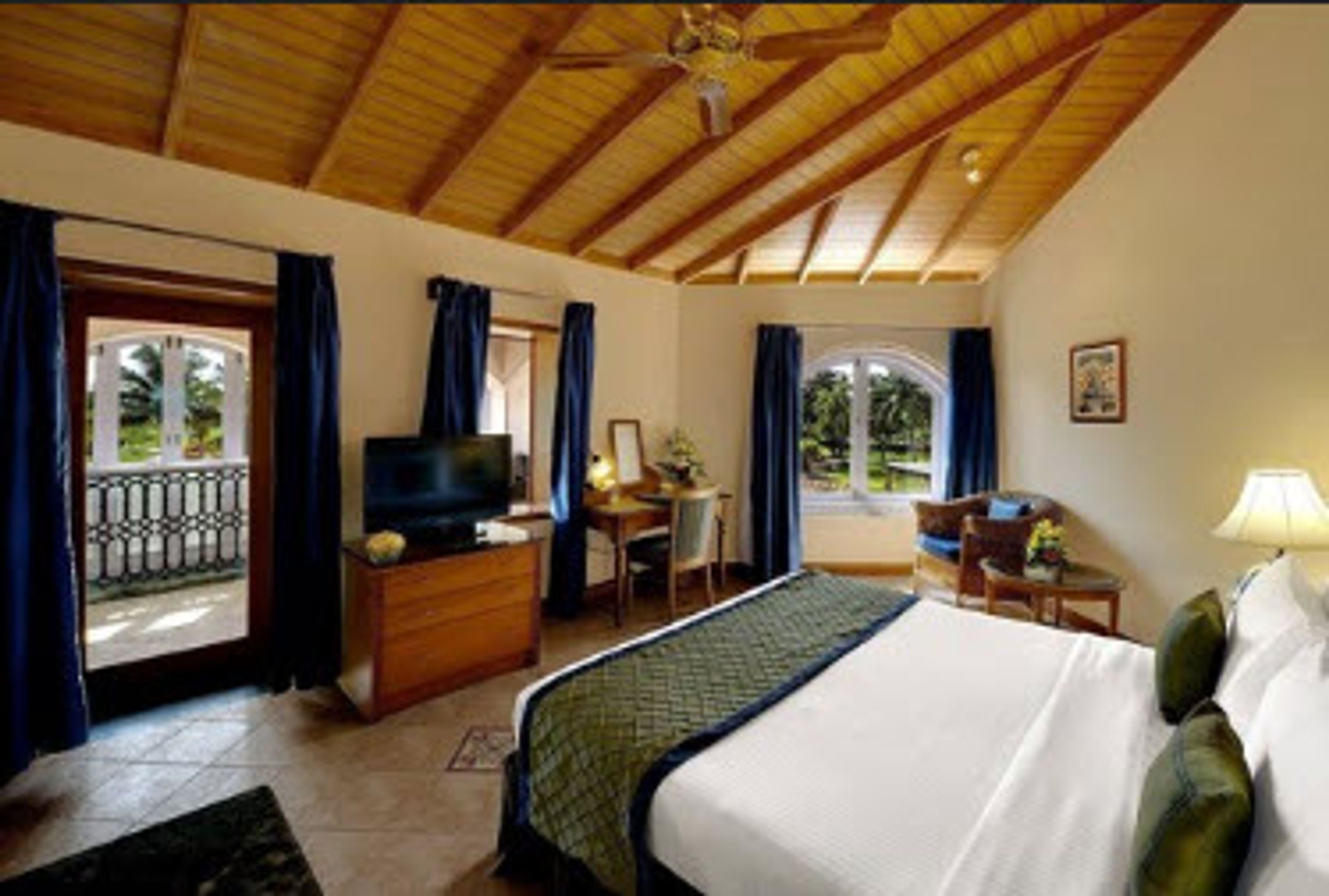 A hotel room with a wooden ceiling, a bed, a desk with a chair, a television, and a window with curtains.