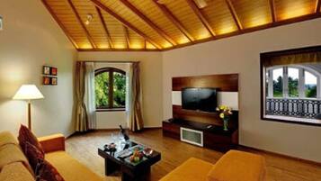 Uma sala de estar com teto de madeira, uma televisão sobre um suporte de madeira, um sofá, uma mesa de centro com frutas e uma janela com cortinas.