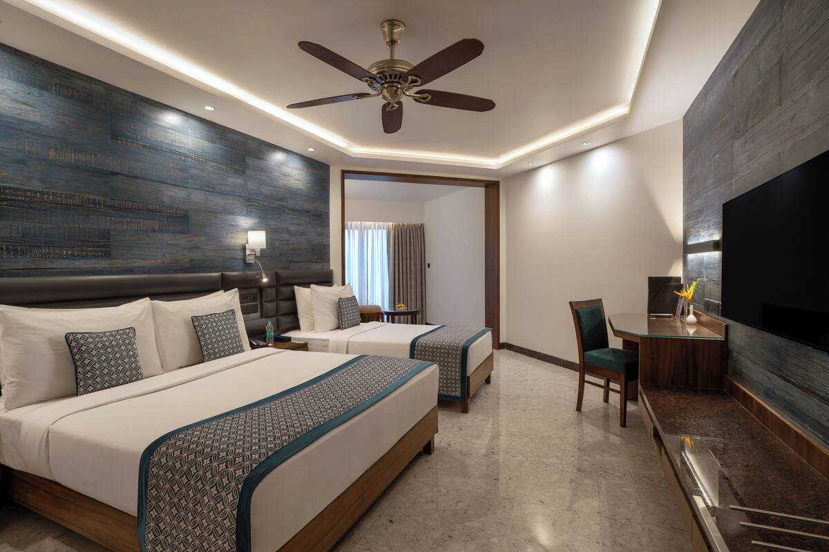 A hotel room with two beds, a desk, a chair, a TV, and a ceiling fan.