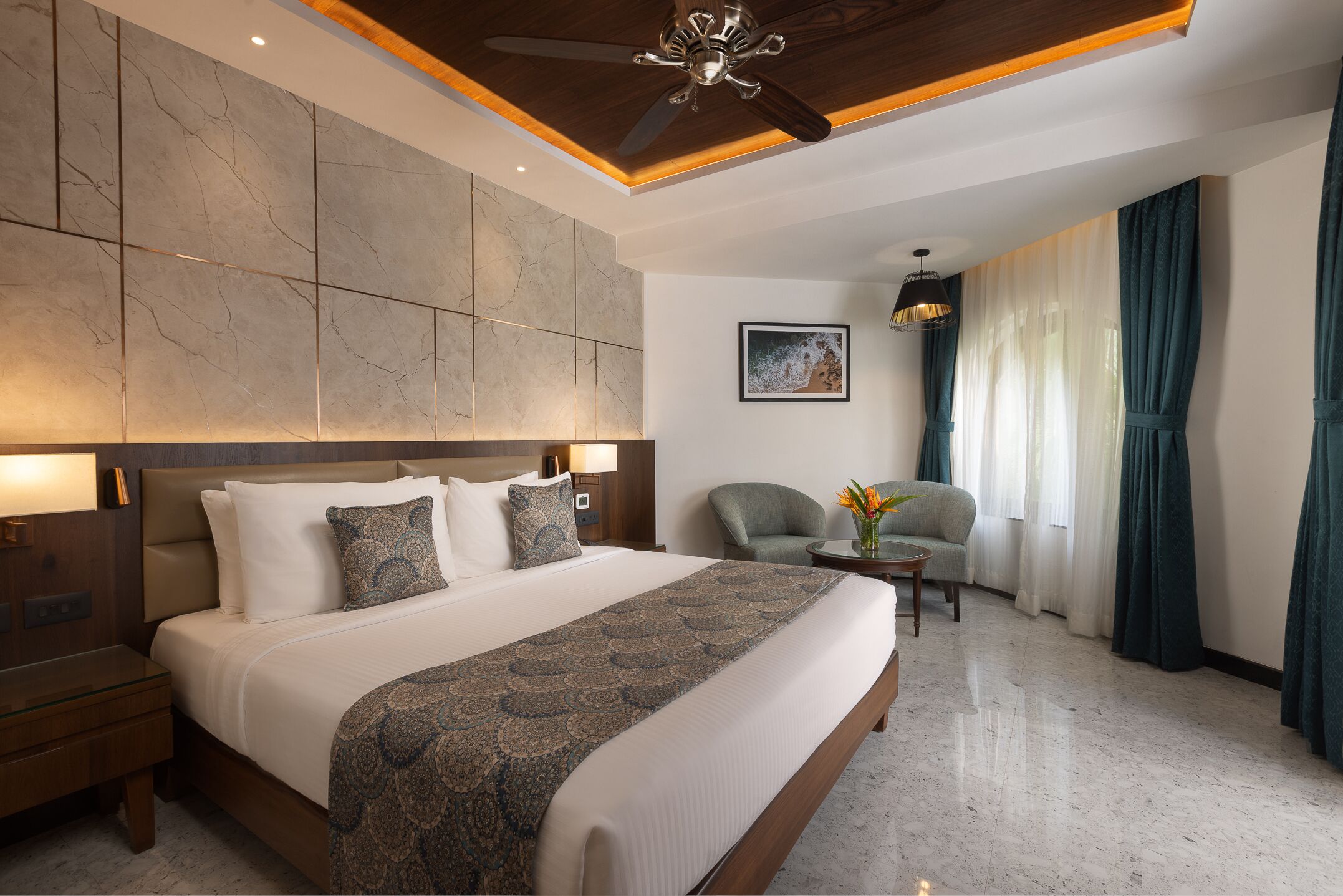 Ein modernes Hotelzimmer mit einem großen Bett, einem Deckenventilator, einer Sitzecke mit einem Sessel und einem kleinen Tisch sowie einem großen Fenster mit Vorhängen.