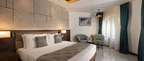 A modern hotel room with a large bed, a ceiling fan, a seating area with a chair and a small table, and a large window with curtains.