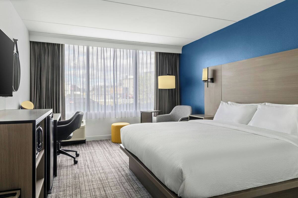 A hotel room with a large bed, a desk with a chair, a small table, a lamp, and a view of the city through the window.
