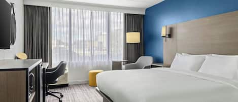 A hotel room with a large bed, a desk with a chair, a small table, a lamp, and a view of the city through the window.