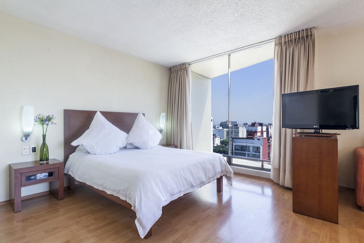 Une chambre d’hôtel avec un lit, un téléviseur posé sur un meuble, une table de chevet avec un vase et une fenêtre avec des rideaux.