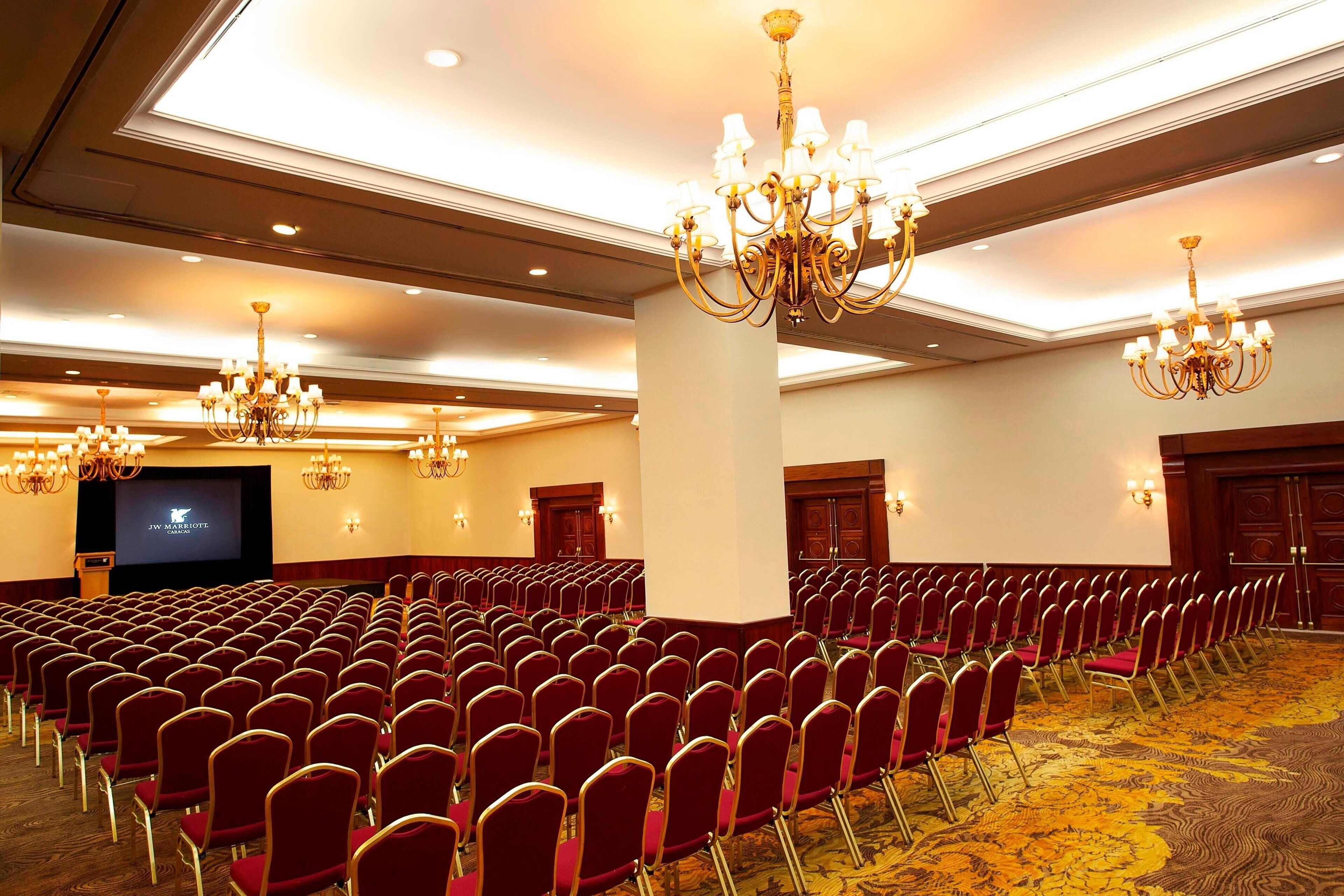 A conference room with rows of red chairs, a large screen, and chandeliers.