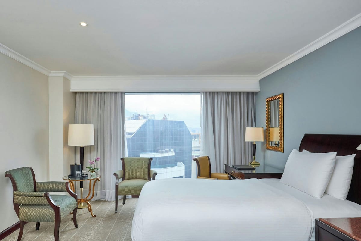 Habitación de hotel con una cama grande, dos sillones, una mesita de noche con lámpara y vistas a la ciudad desde la ventana.