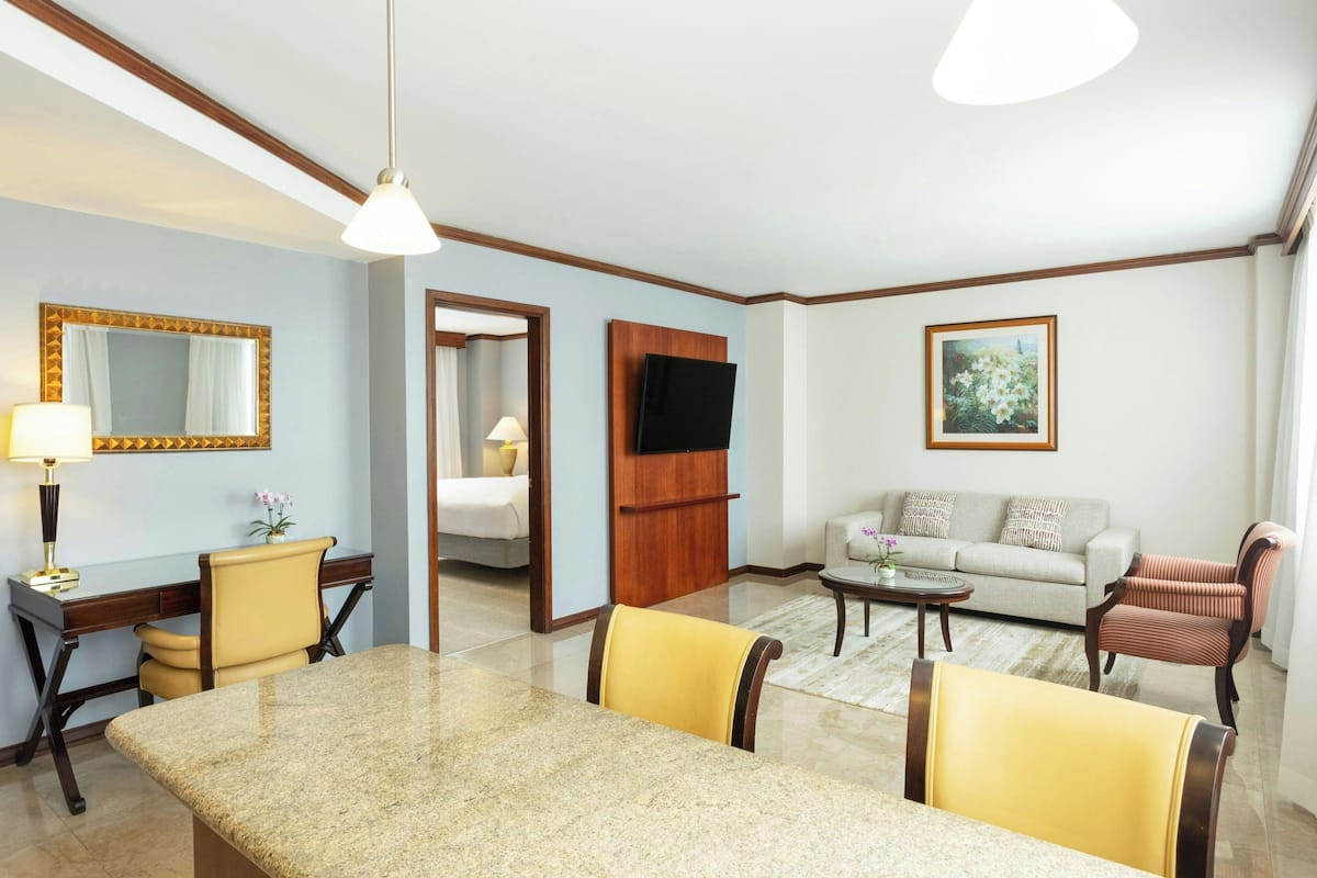 Una habitación de hotel con zona de comedor, un sofá, un televisor y un cuadro en la pared.