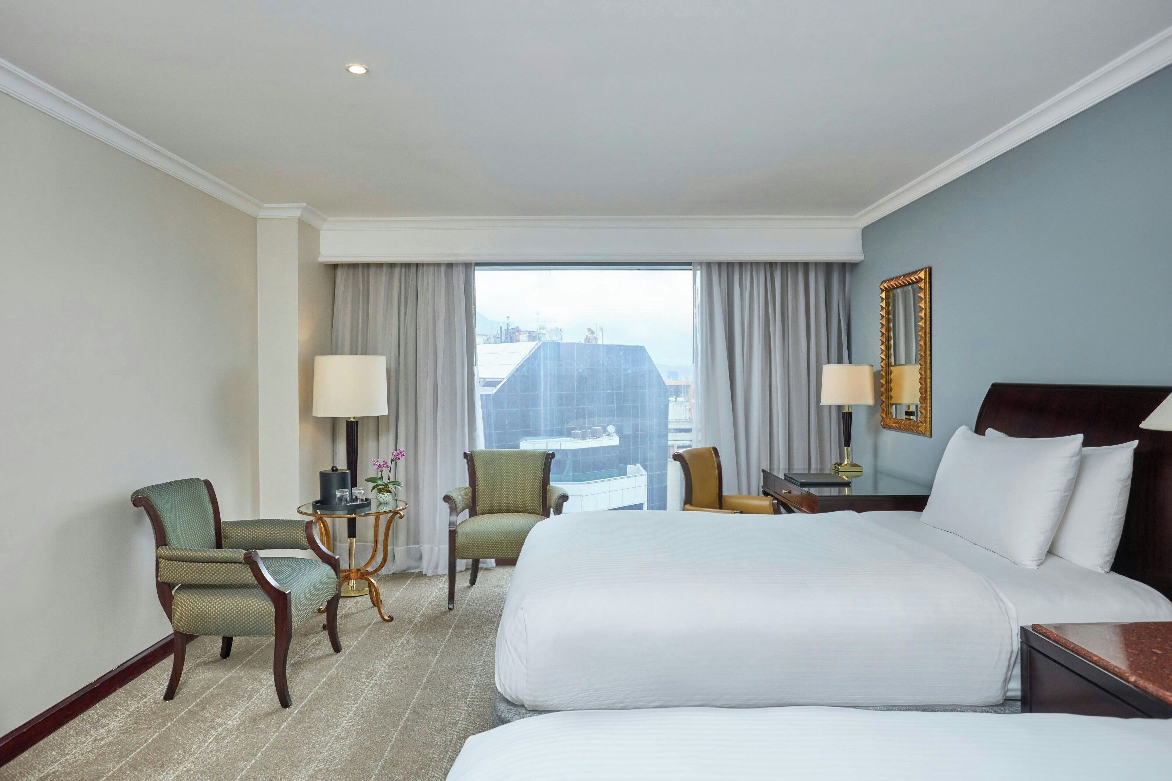 A hotel room with a large bed, two chairs, a small table, a lamp, and a view of the city through the window.