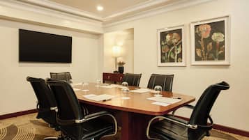 A conference room with a large wooden table, black leather chairs, a flat-screen TV, framed floral artwork, and a wall-mounted lamp.