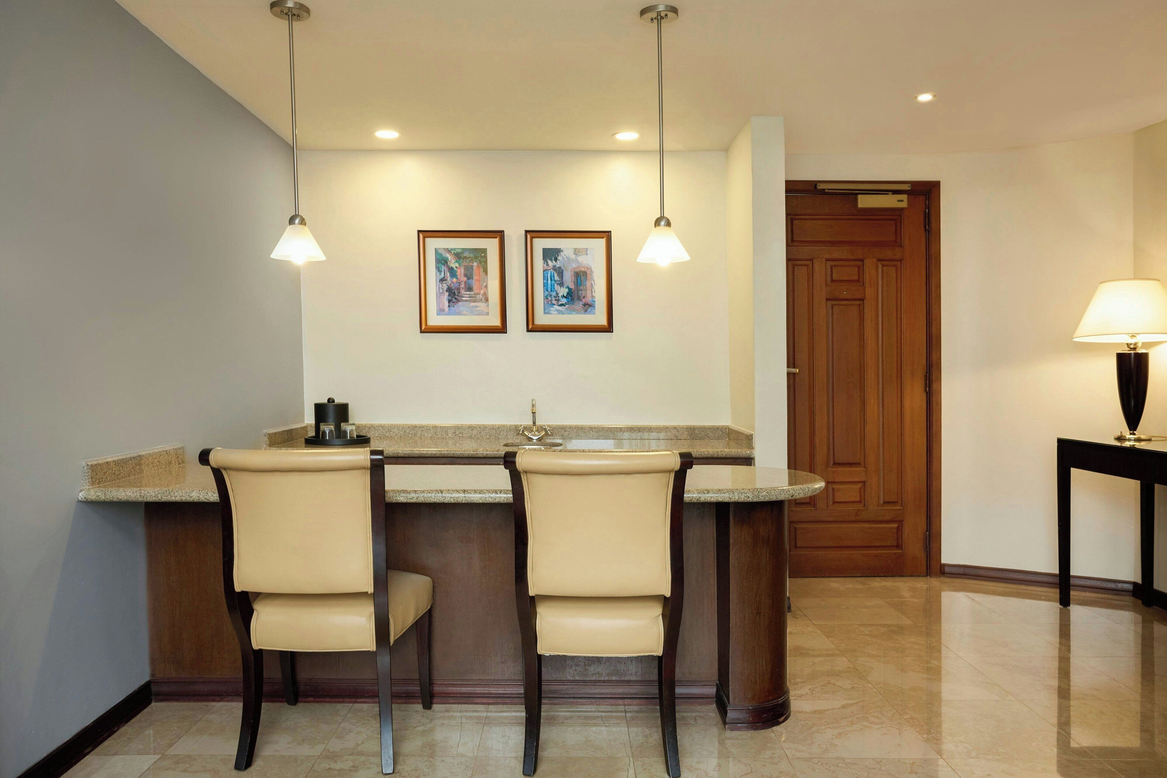 A kitchen with a bar area, two chairs, a wooden door, and a table with a lamp.