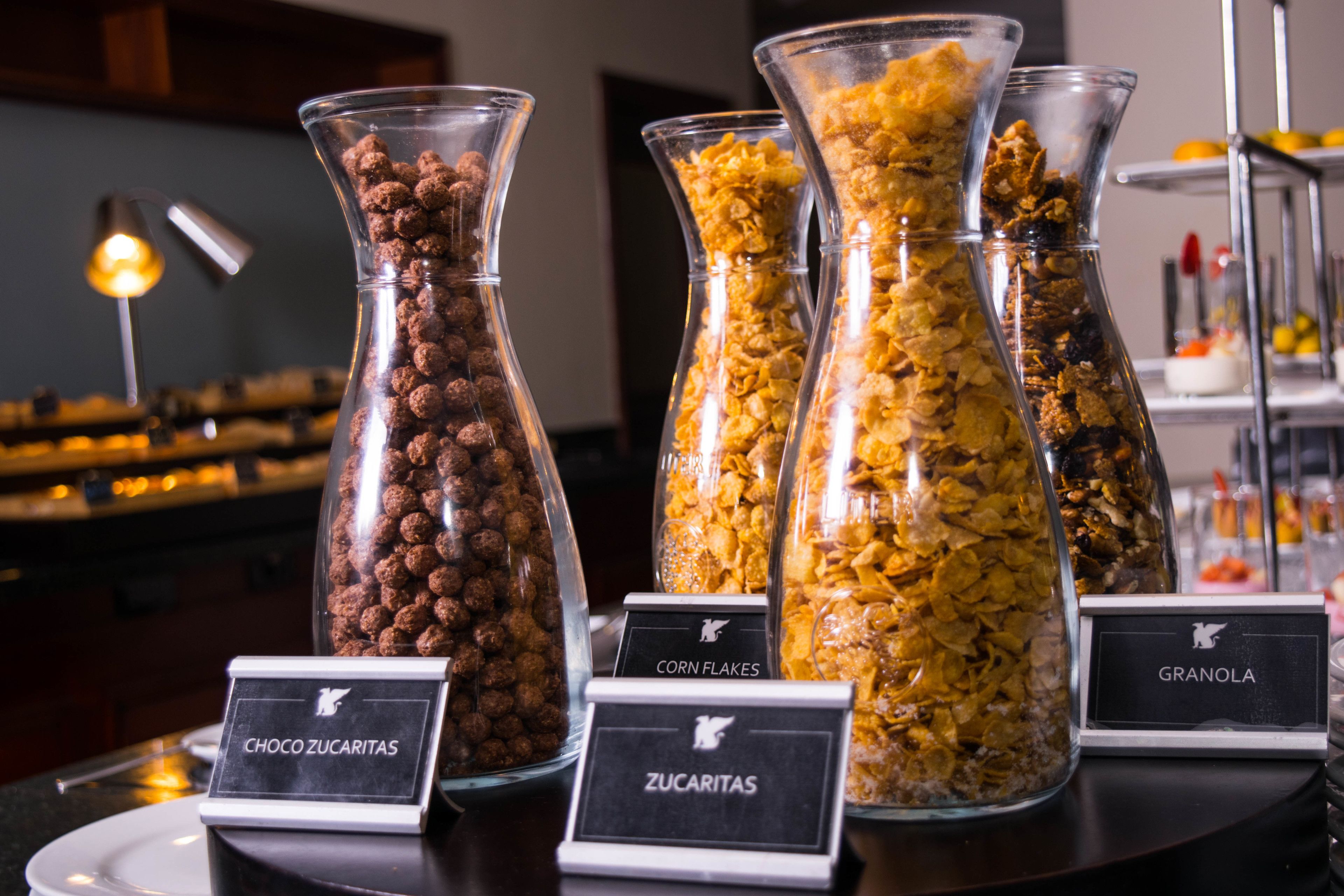 A buffet with clear glass containers labeled with different types of snacks like chocozucaritas, corn flakes, and granola.