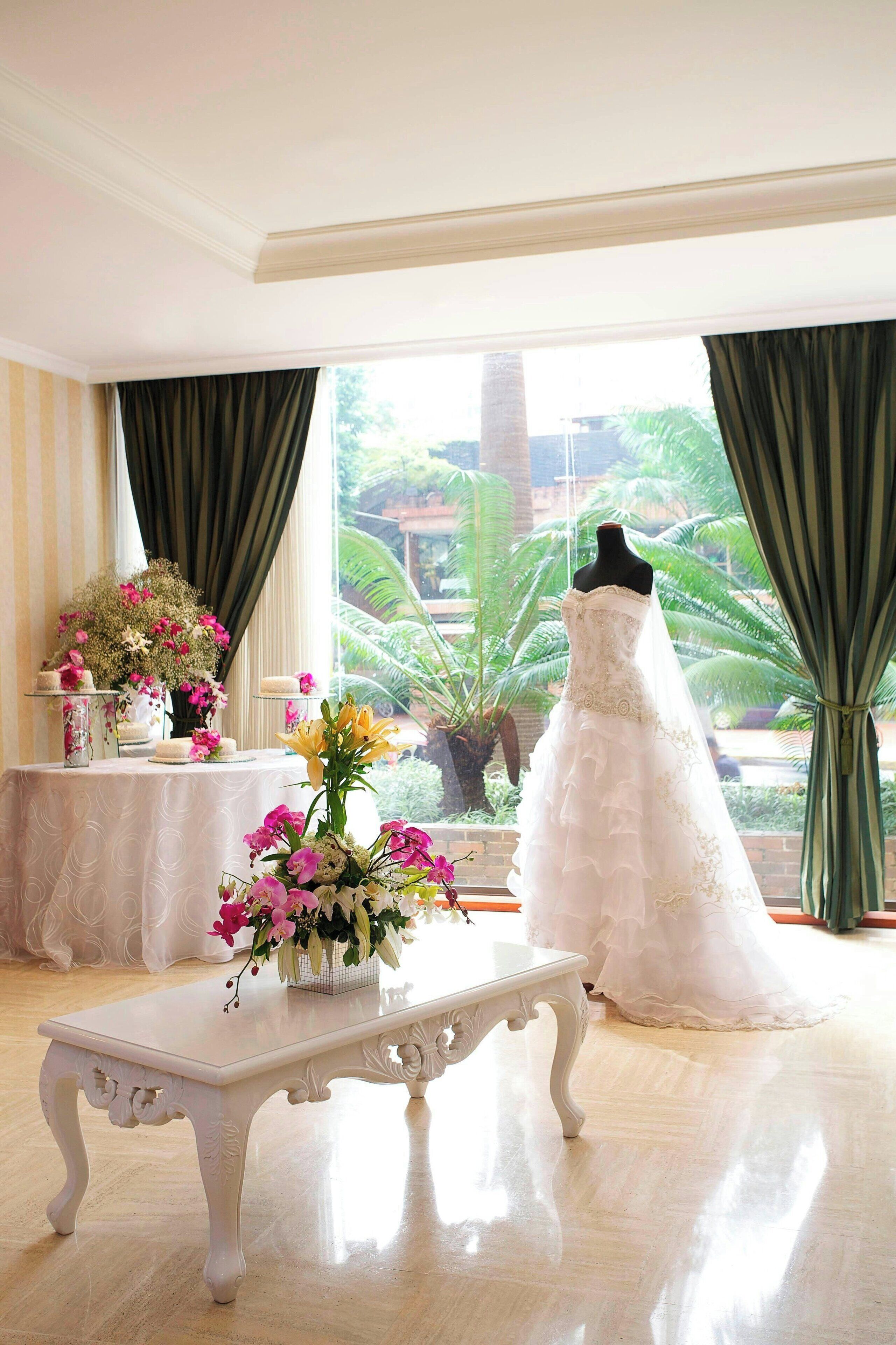 A room with a mannequin in a wedding dress, a table with floral arrangements, and a view of greenery outside.