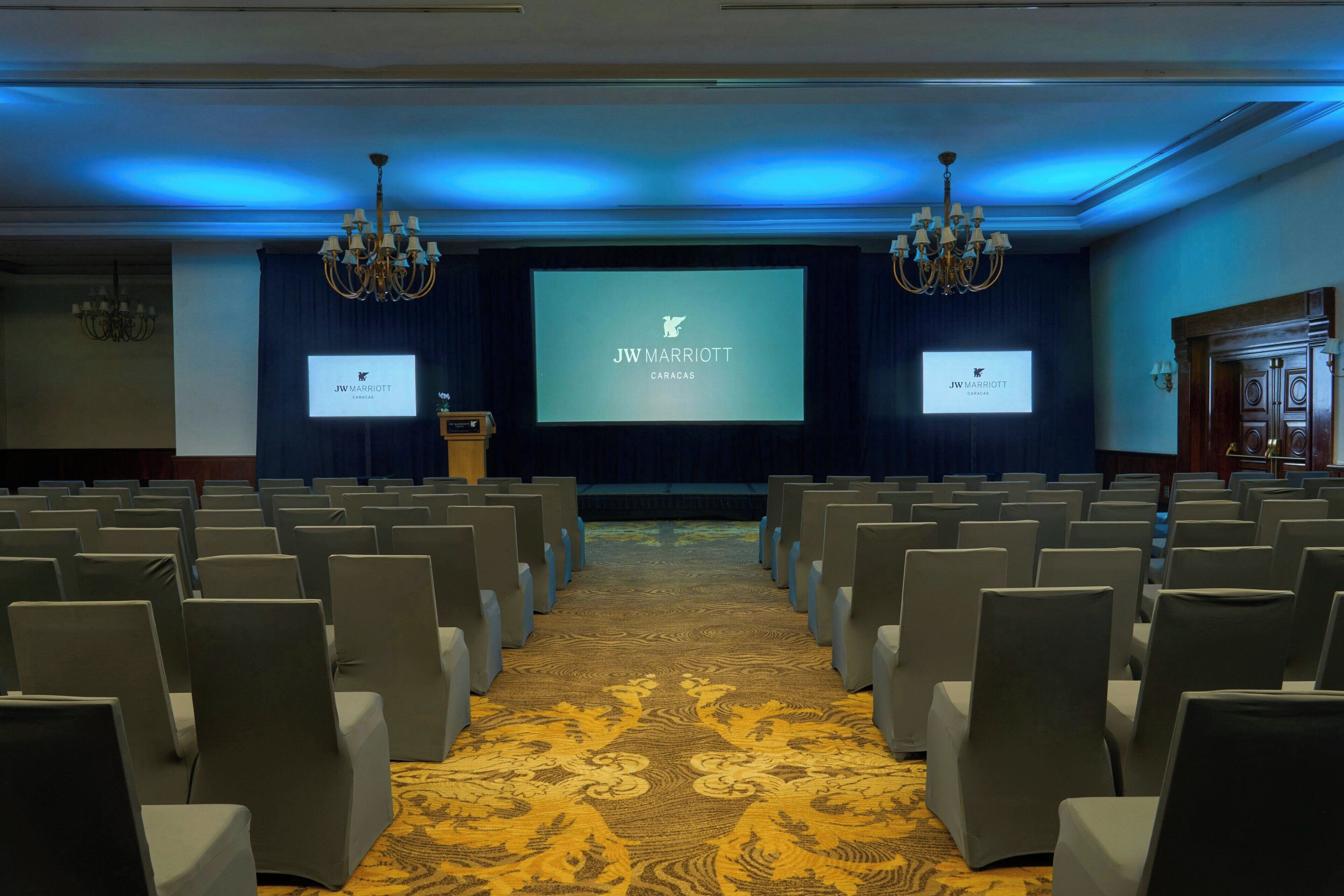 A conference room with rows of chairs, a stage with a screen displaying 'JW MARRIOTT CARACAS', chandeliers, and a patterned carpet.