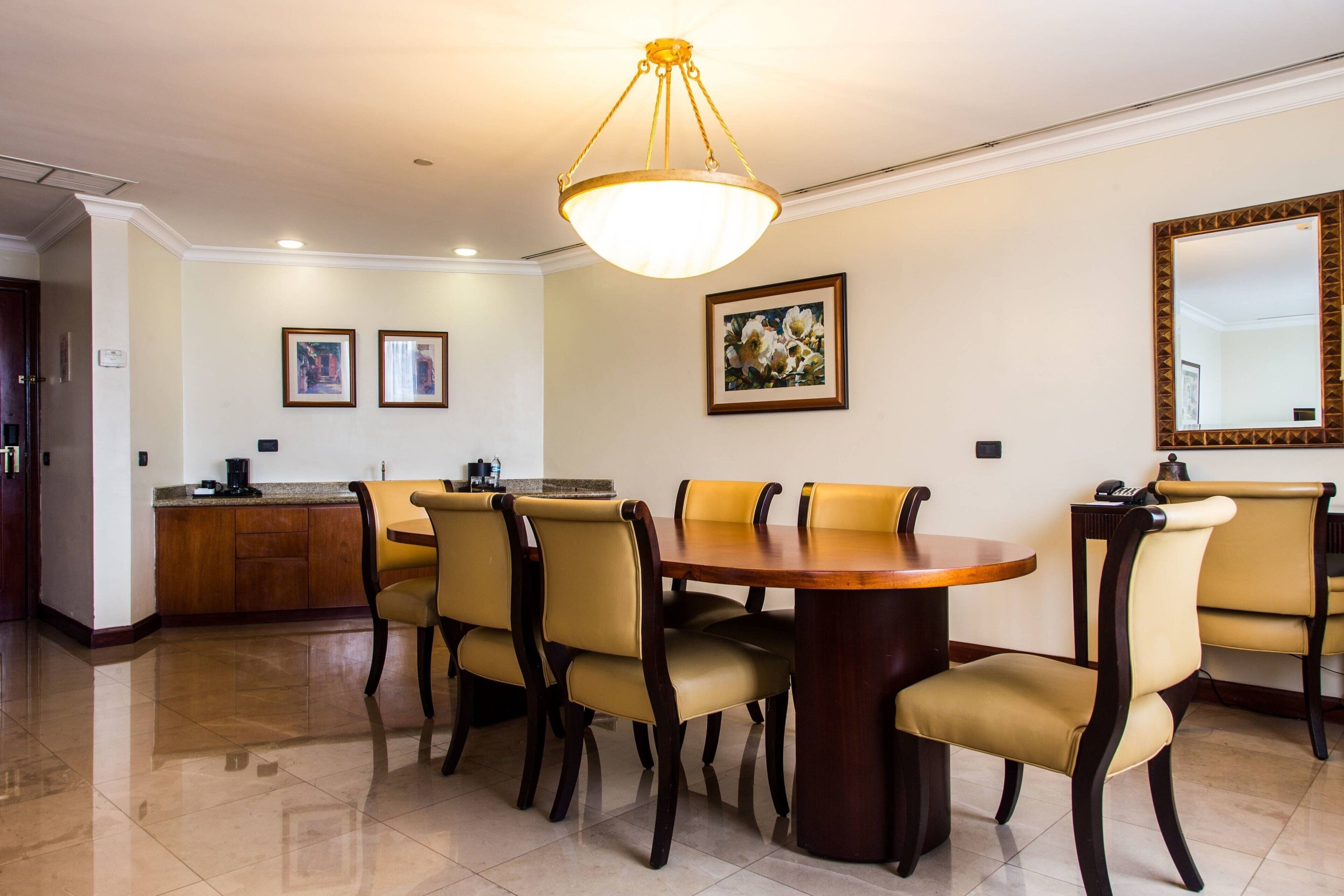 A dining area with a round table and chairs, a kitchen in the background, and a large mirror on the wall.