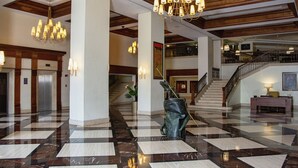 Un vestíbulo amplio con un piso de mármol a cuadros, una escultura central y una escalera que conduce a un nivel superior.