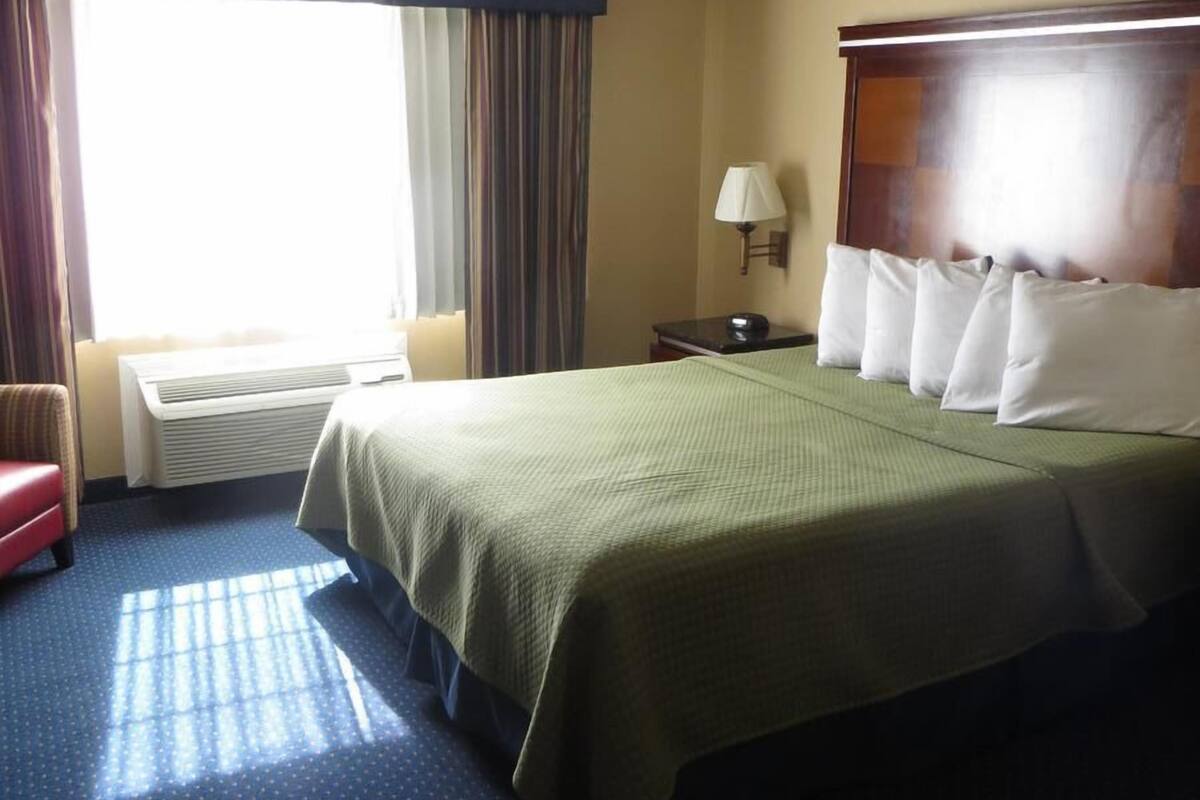 Ein Hotelzimmer mit einem großen Bett, einem Nachttisch, einer Lampe, einem Sessel und einem Fenster mit Vorhängen.