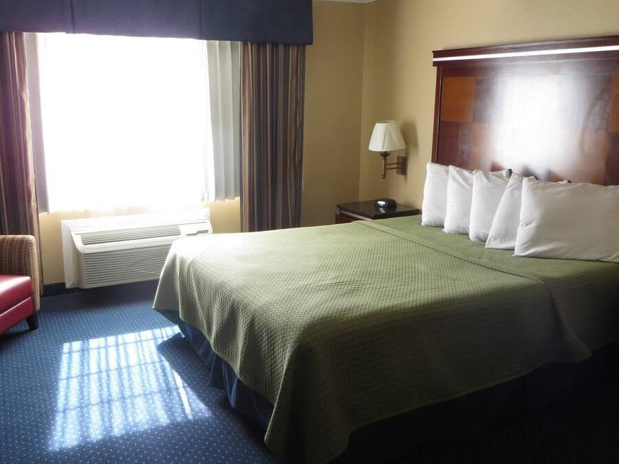 Ein Hotelzimmer mit Bett, Nachttisch, Lampe, Stuhl und einem fenster mit Vorhängen.