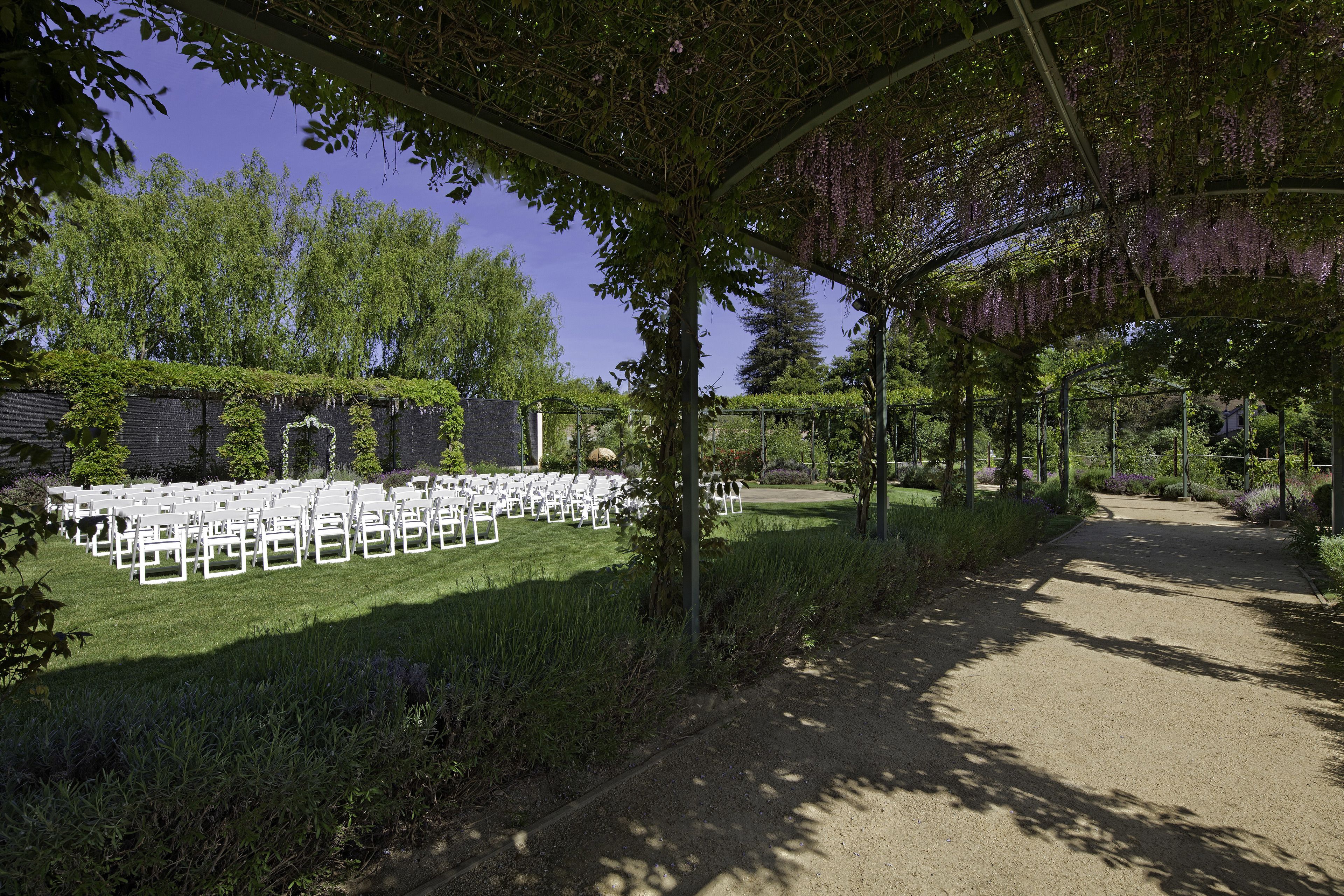 A covered outdoor seating area with white chairs arranged in rows, surrounded by greenery and trees.
