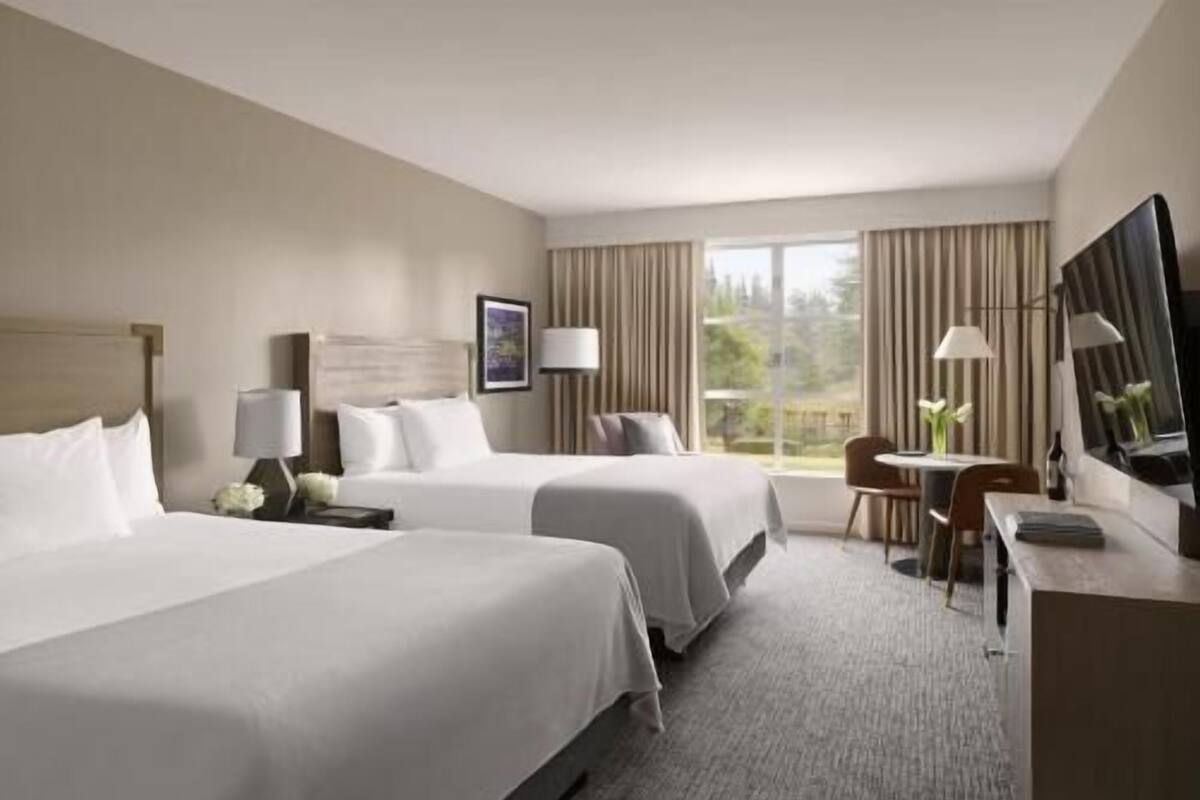 A hotel room with two beds, a desk, a chair, a TV, and a window with curtains.