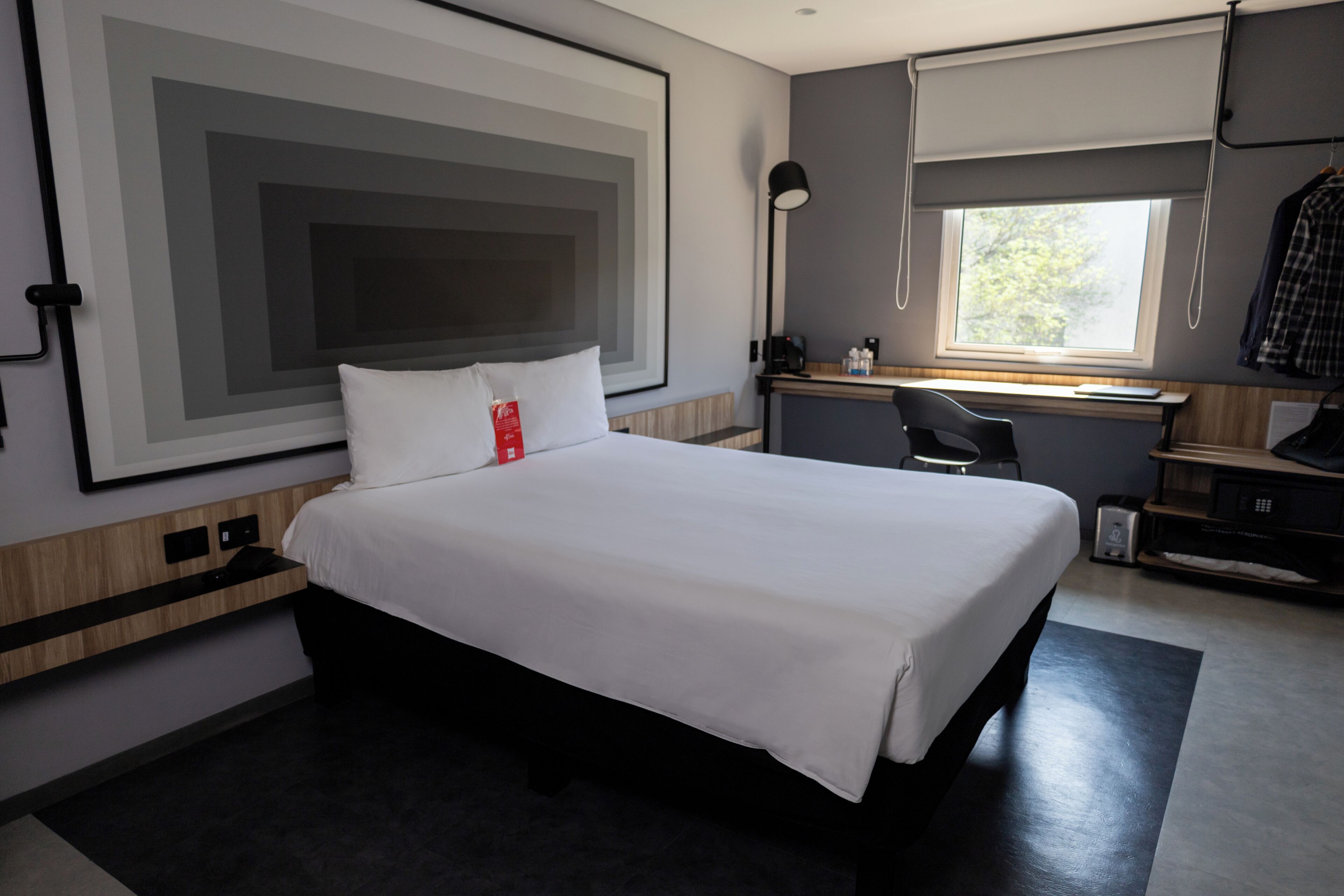 A modern hotel room with a large bed, a desk, a chair, a lamp, and a window.