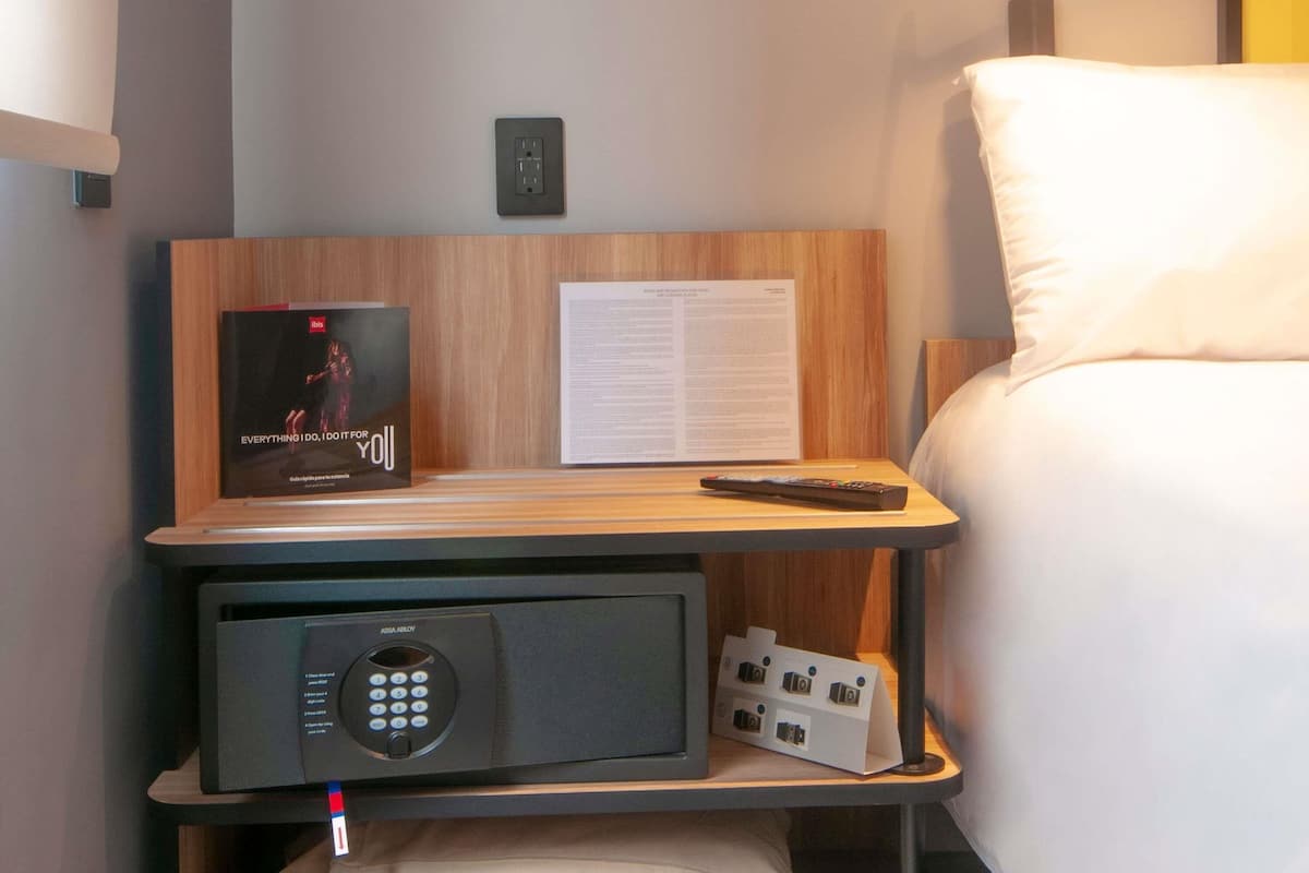 Habitación de hotel ordenada con una mesita de noche que sostiene un control remoto, una revista y un pequeño dispositivo electrónico.
